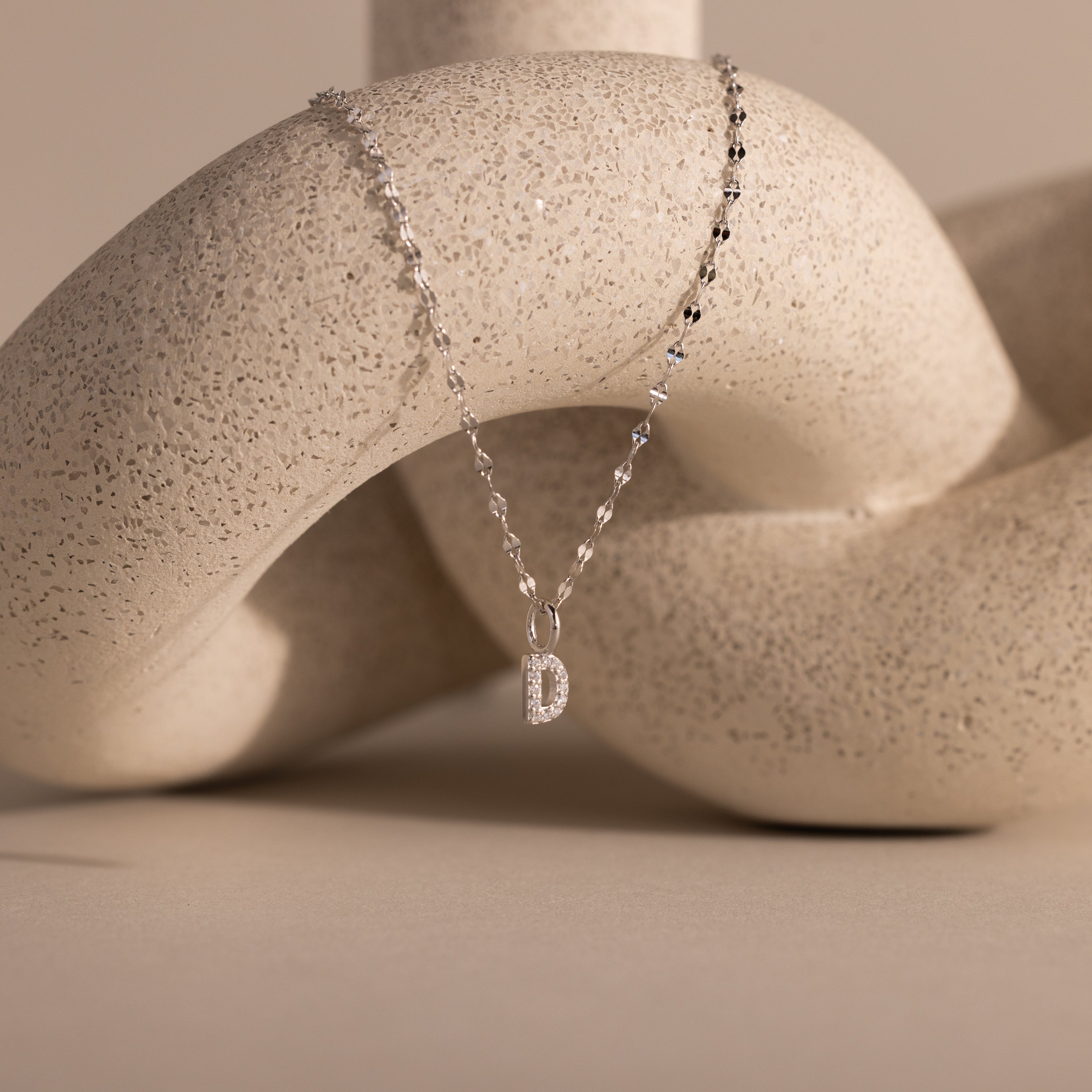 The Dainty Pave Letter Necklace in Sterling Silver, featuring a delicate pavé letter D pendant, drapes elegantly over a textured abstract beige sculpture.