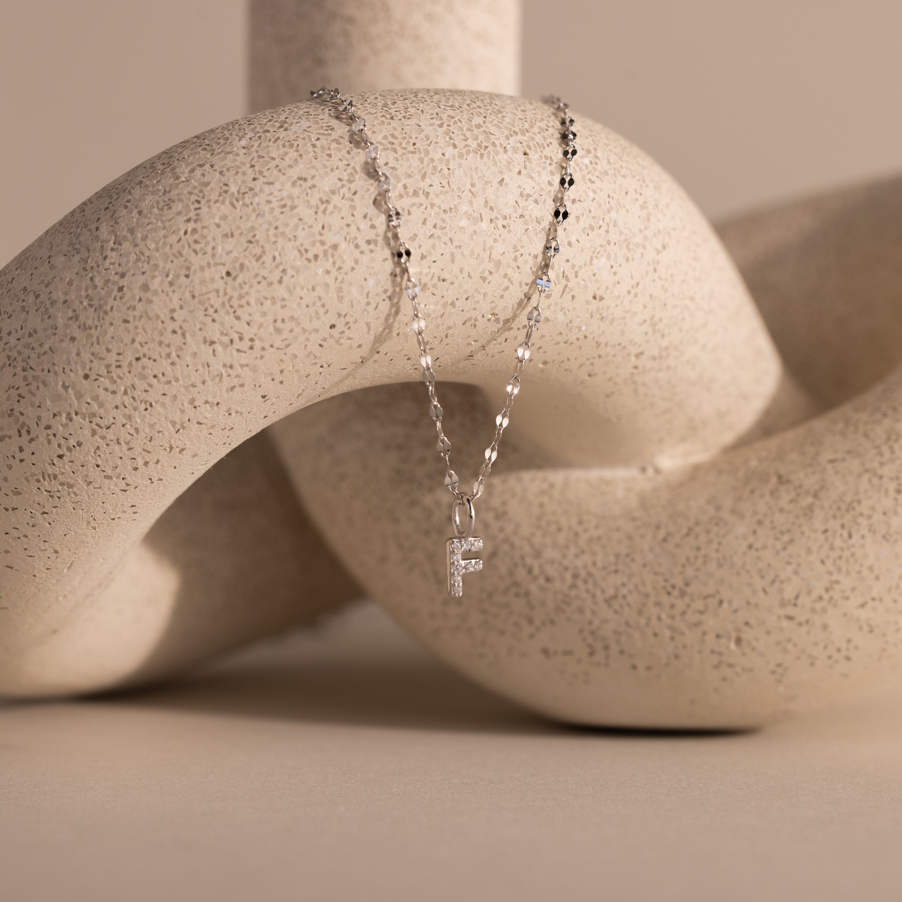 The Dainty Pave Letter Necklace in Sterling Silver, featuring an F pendant, sparkles like a CZ diamond as it drapes elegantly over a textured beige abstract sculpture.