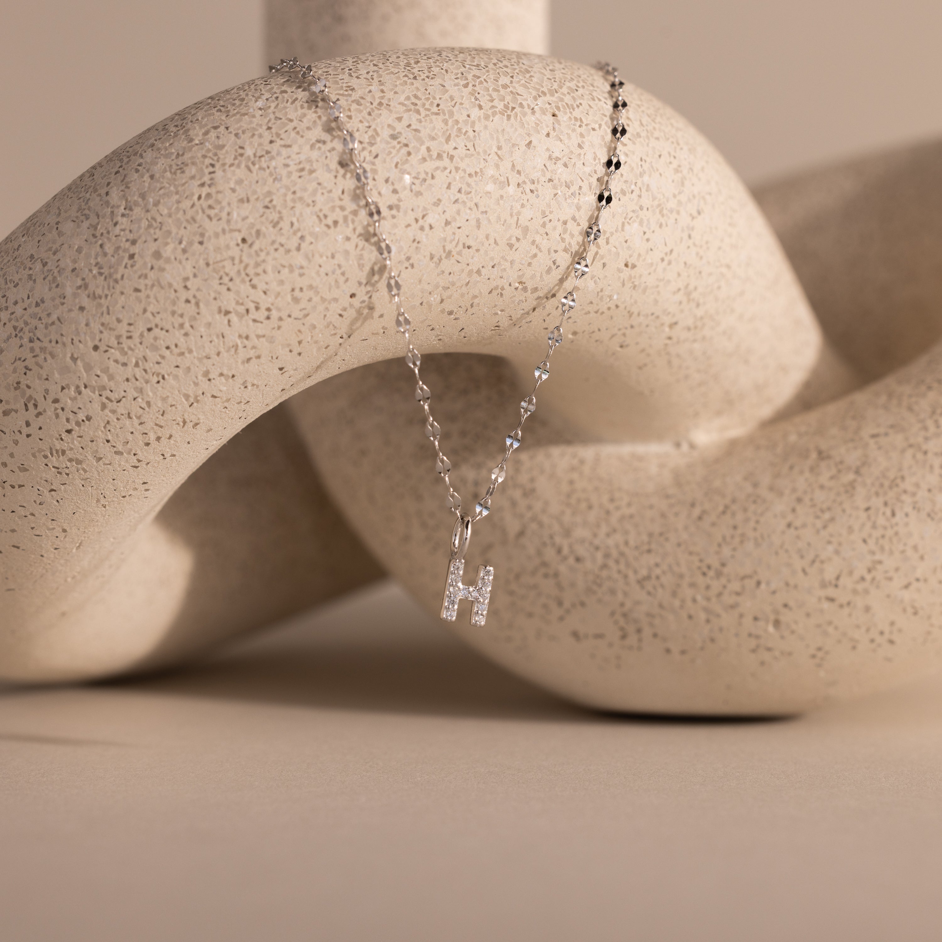 The Dainty Pave Letter Necklace in Sterling Silver features an H pendant and is shown draped over a textured beige abstract sculpture.