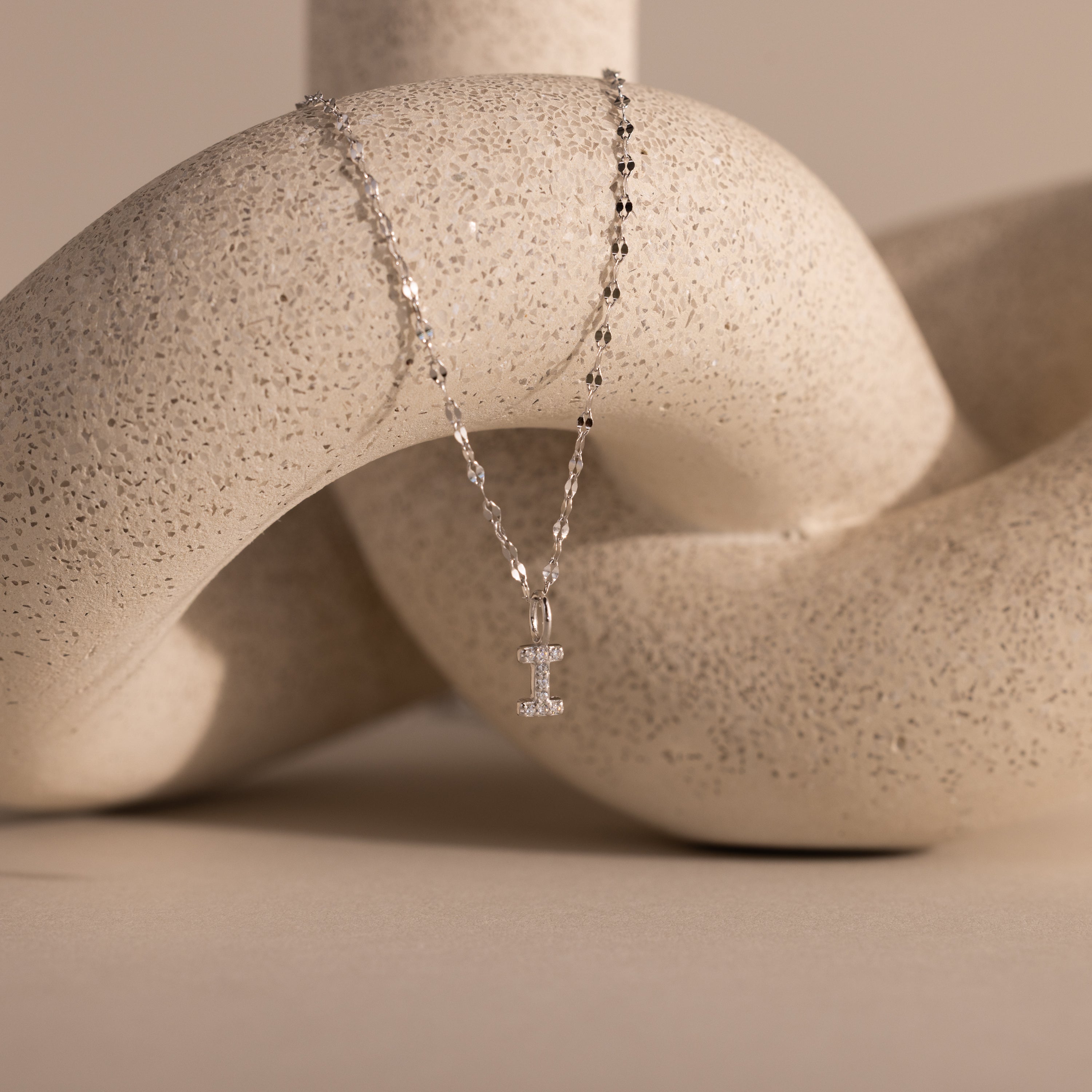 The Dainty Pave Letter Necklace in Sterling Silver, featuring a sparkling I pendant, is displayed on a textured, abstract beige sculpture.