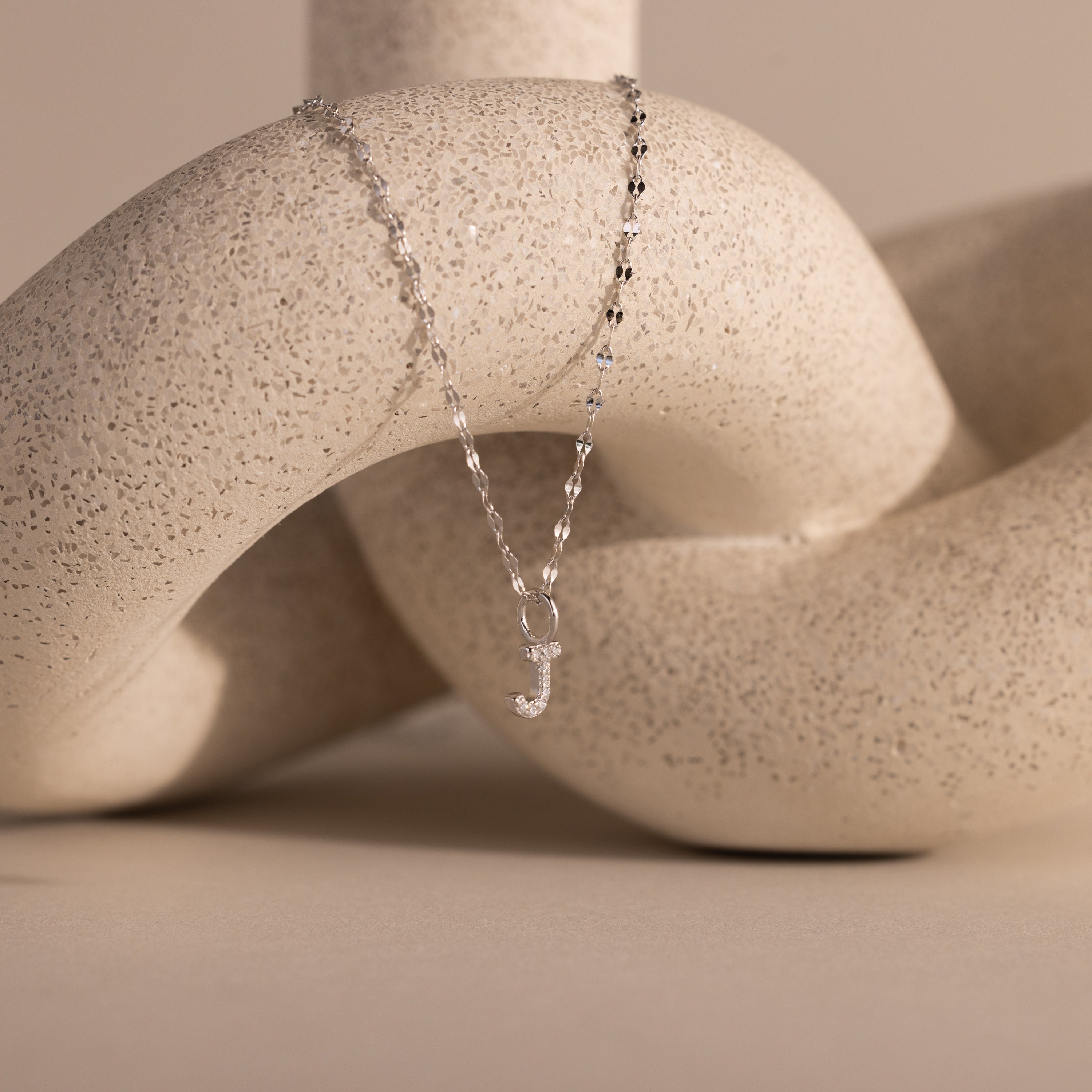 The Dainty Pave Letter Necklace in Sterling Silver features a shimmering pavé letter pendant elegantly displayed over a textured, intertwined ceramic sculpture.