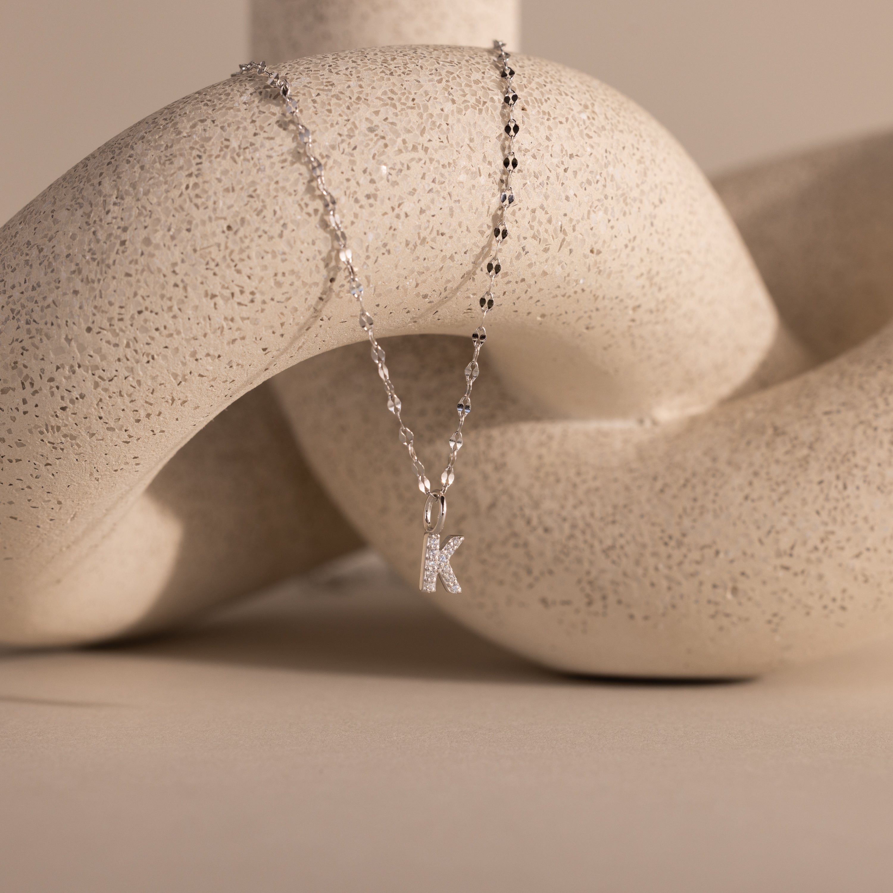 The Dainty Pave Letter Necklace in Sterling Silver, featuring a sparkling K pendant, drapes elegantly over a textured, abstract cream-colored sculpture.