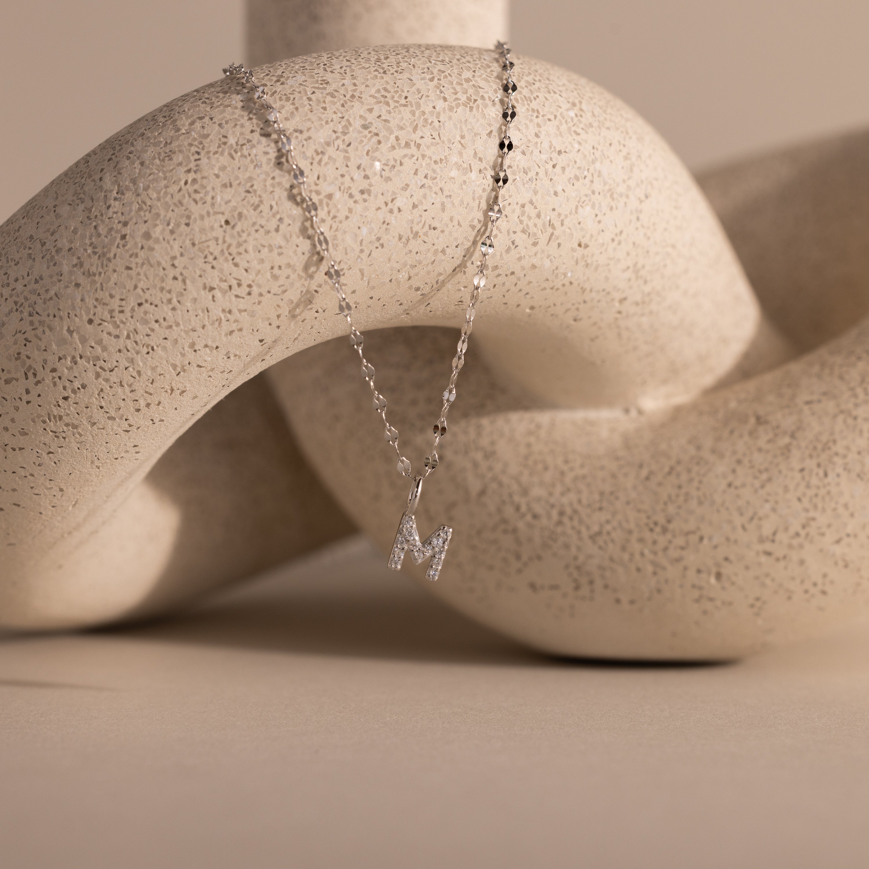 The Dainty Pave Letter Necklace in Sterling Silver, featuring a sparkling CZ M pendant, drapes elegantly over a textured white sculpture.