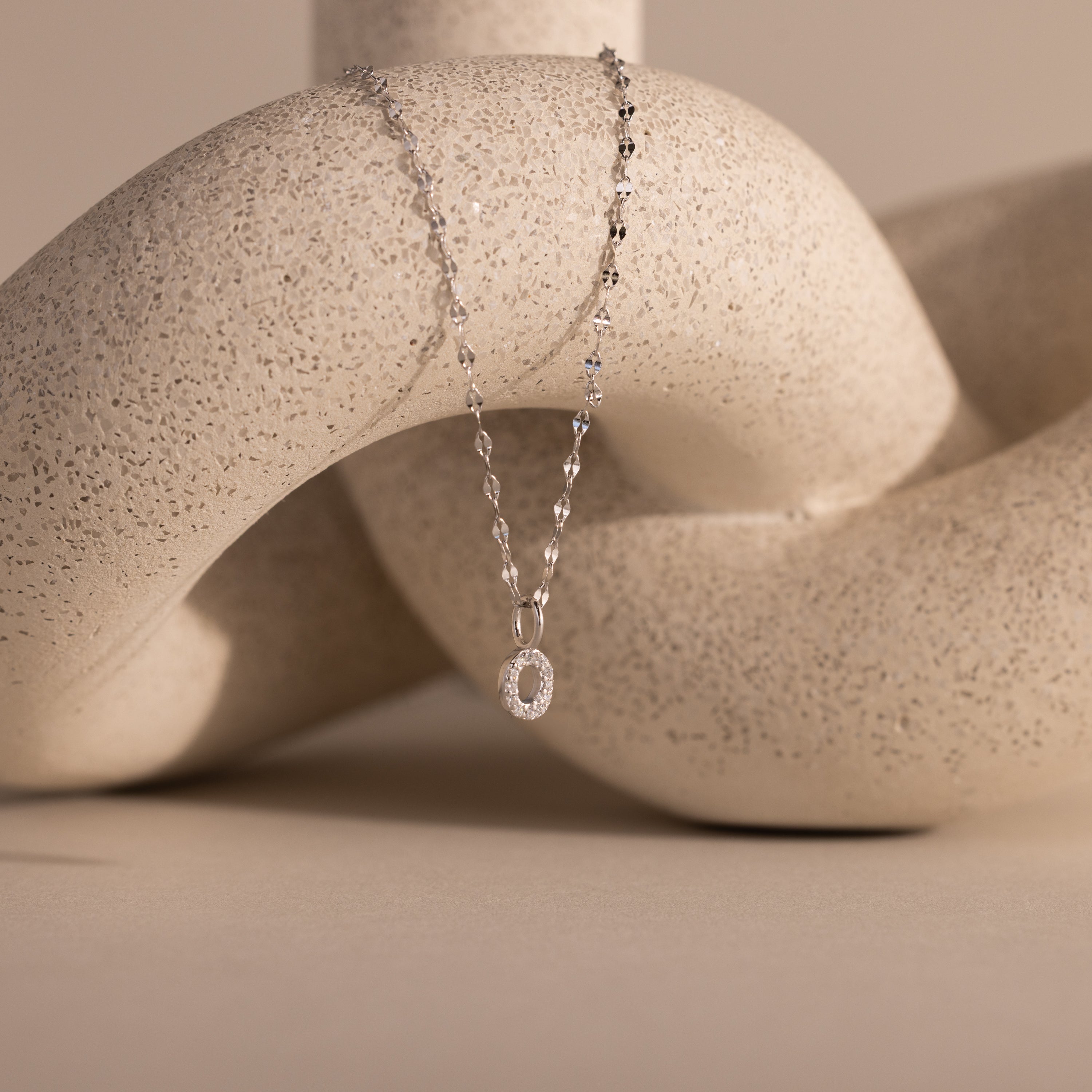 The Dainty Pave Letter Necklace in Sterling Silver, featuring a delicate silver pendant, is elegantly draped over a light, textured abstract sculpture.