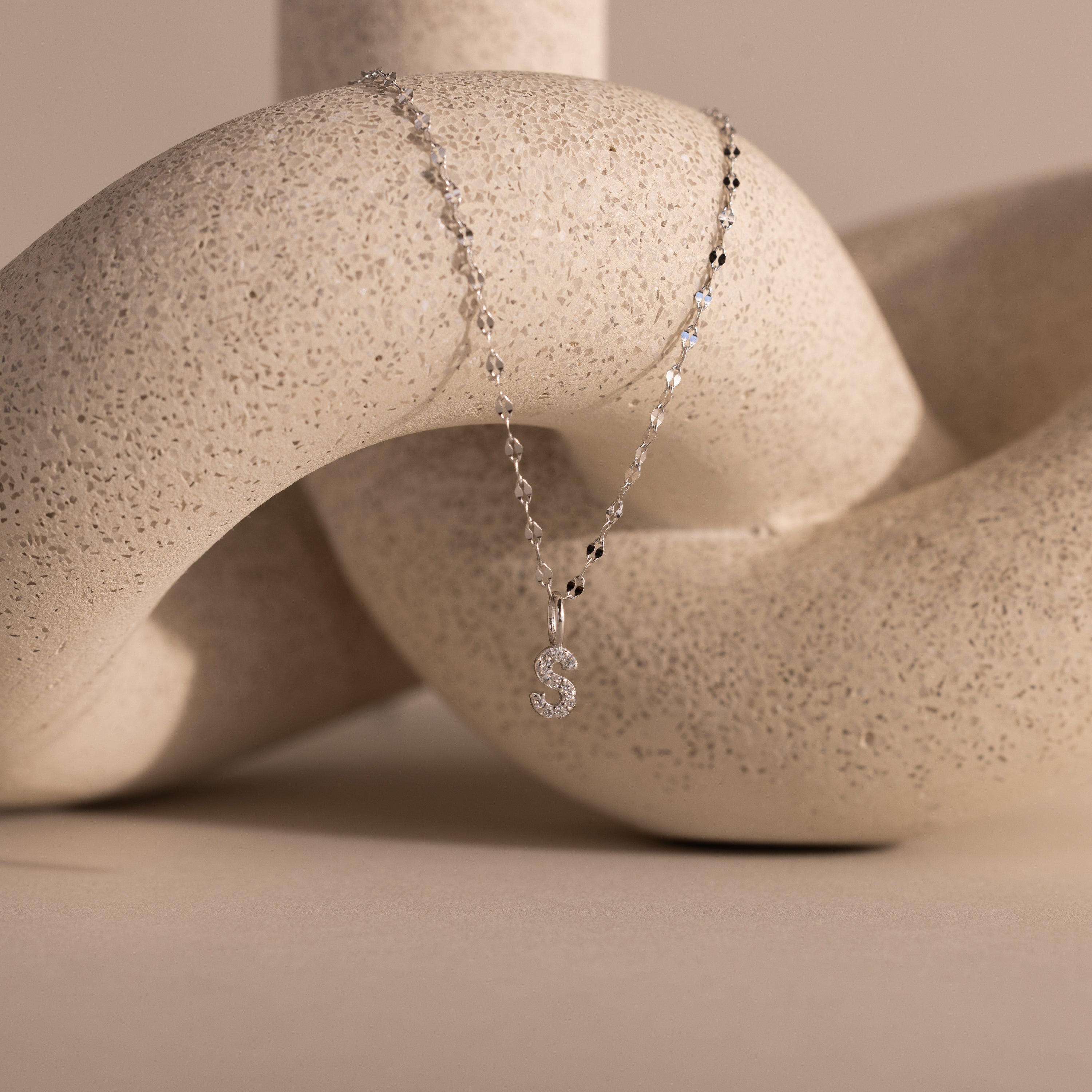 Dainty Pave Letter Necklace in Sterling Silver with an S pendant beautifully draped over a textured, curved, beige sculpture.