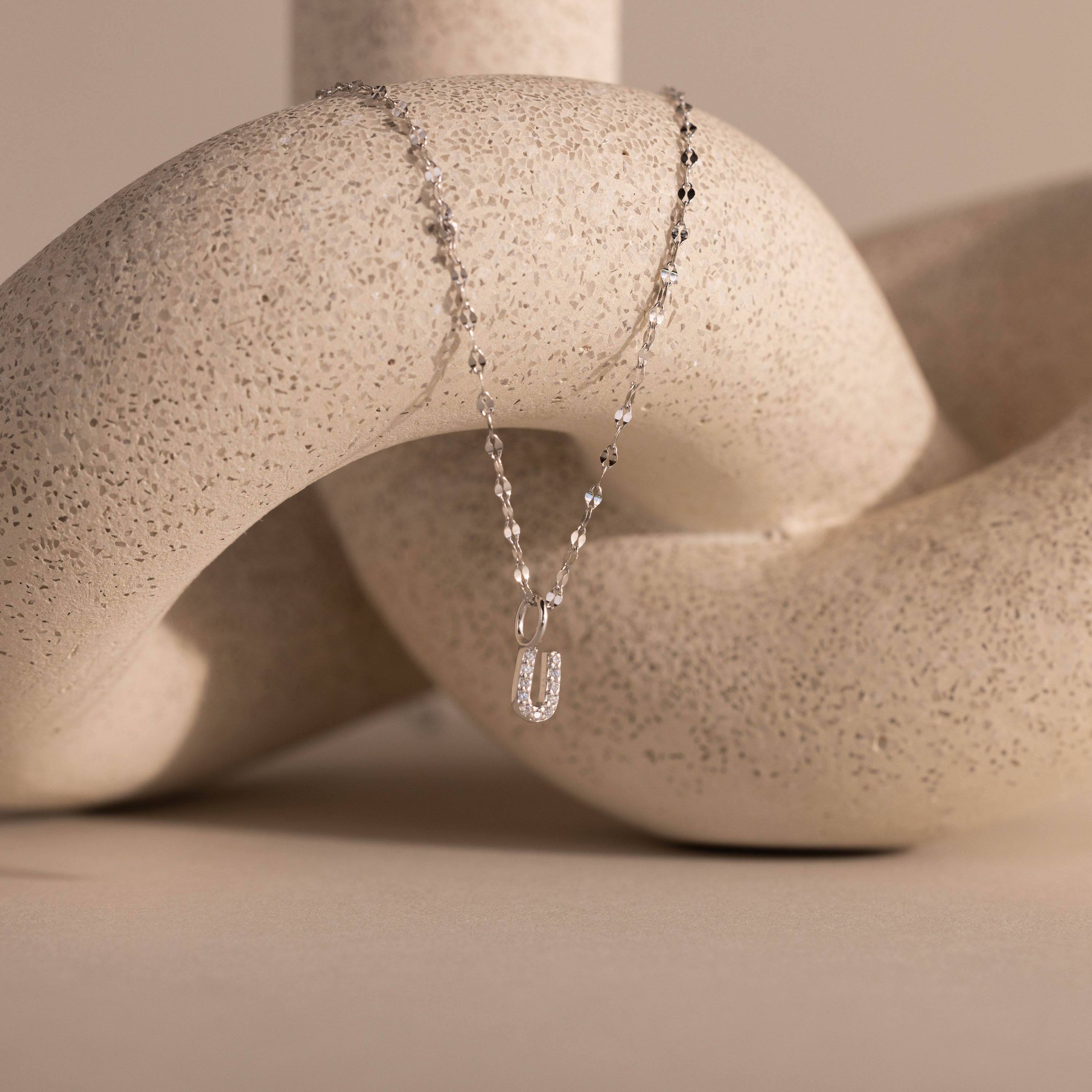 The Dainty Pave Letter Necklace in Sterling Silver, featuring a U-shaped pendant, is elegantly displayed draped over a textured, cream-colored abstract sculpture.