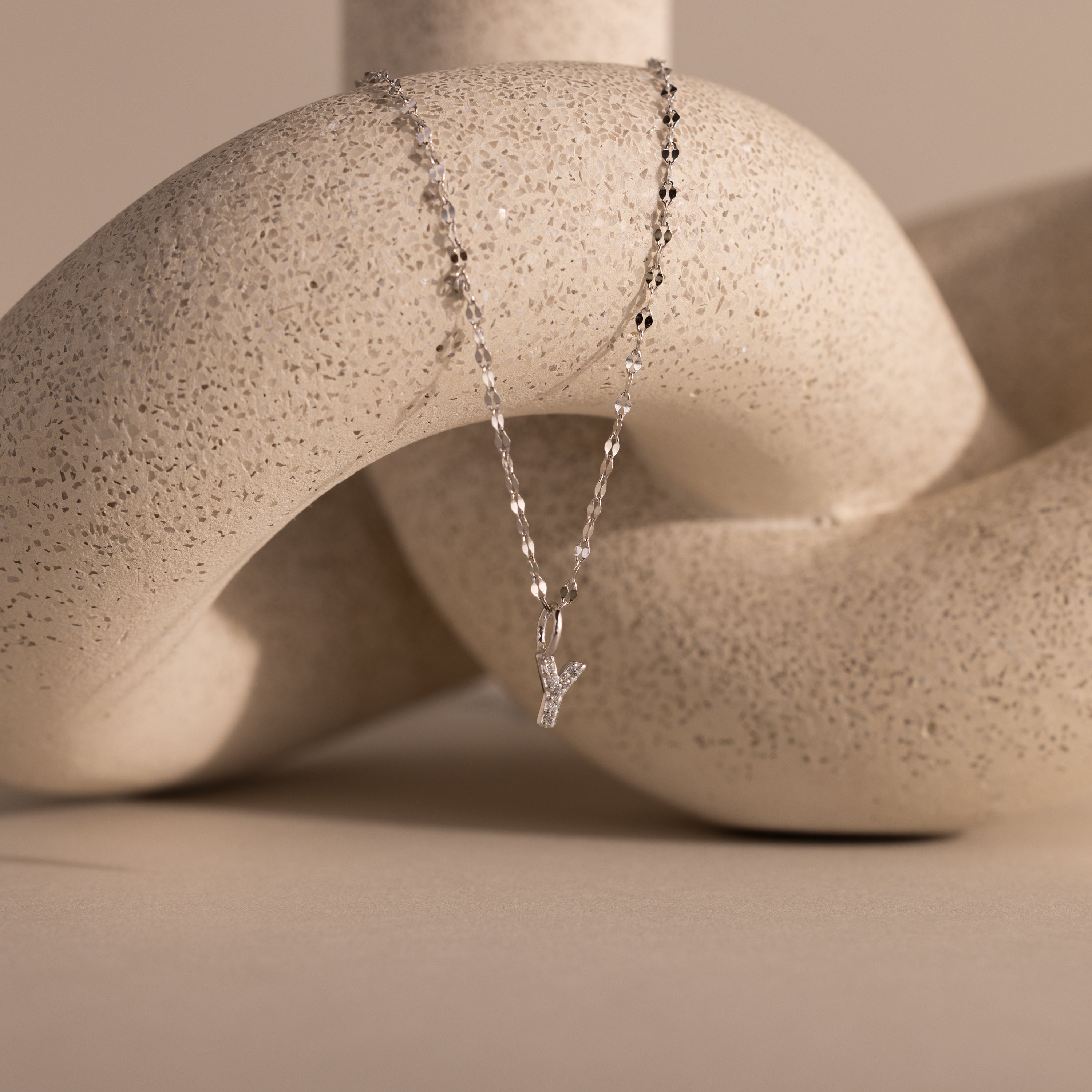 The Dainty Pave Letter Necklace in Sterling Silver is draped over a textured, abstract beige sculpture.