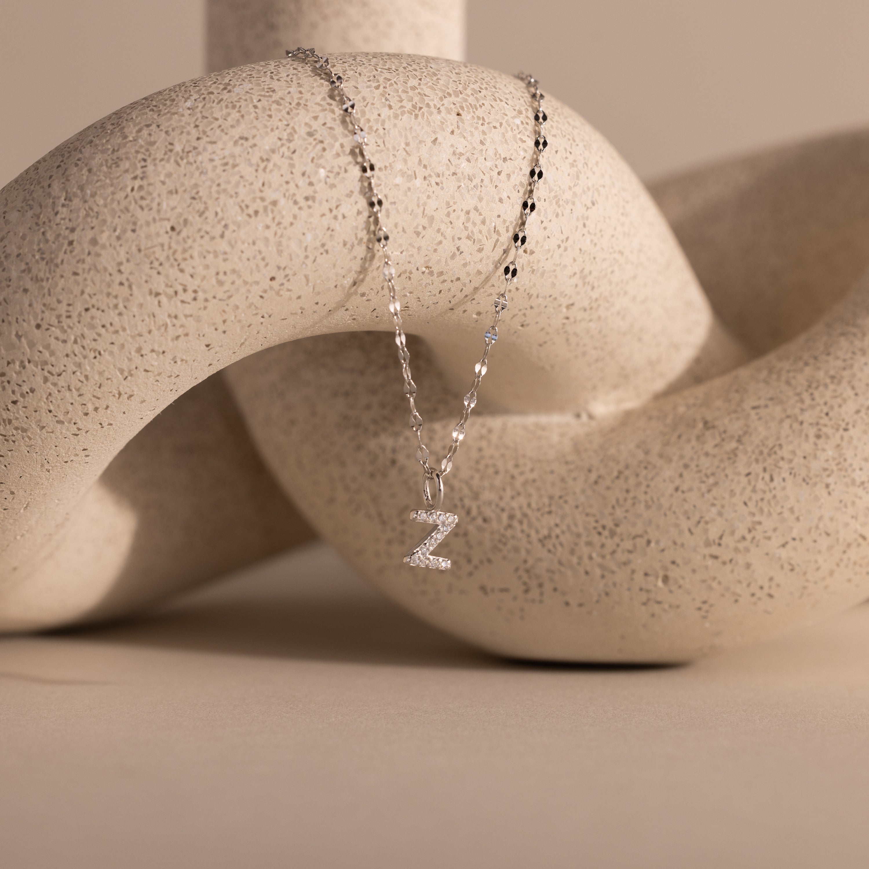The Dainty Pave Letter Necklace in Sterling Silver, featuring a sparkling pavé letter Z pendant, is elegantly displayed draped over a textured, abstract beige sculpture.