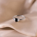 Black Baguette Diamond Ring in Sterling Silver featuring a baguette-cut centerpiece and sparkling clear gems, elegantly displayed on soft beige fabric for a vintage-inspired style.
