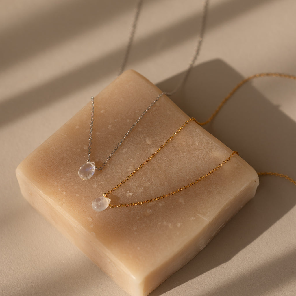 The Teardrop Moonstone Necklace, a delicate minimalist jewelry piece with a pendant, rests on a beige soap bar illuminated by soft, natural light and gentle shadows.