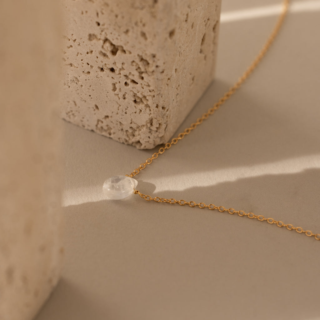The Teardrop Moonstone Necklace in 18K Gold, featuring a clear stone pendant, rests on a beige surface next to textured stone blocks.