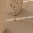 The Teardrop Moonstone Necklace in 18K Gold, featuring a clear stone pendant, rests on a beige surface next to textured stone blocks.