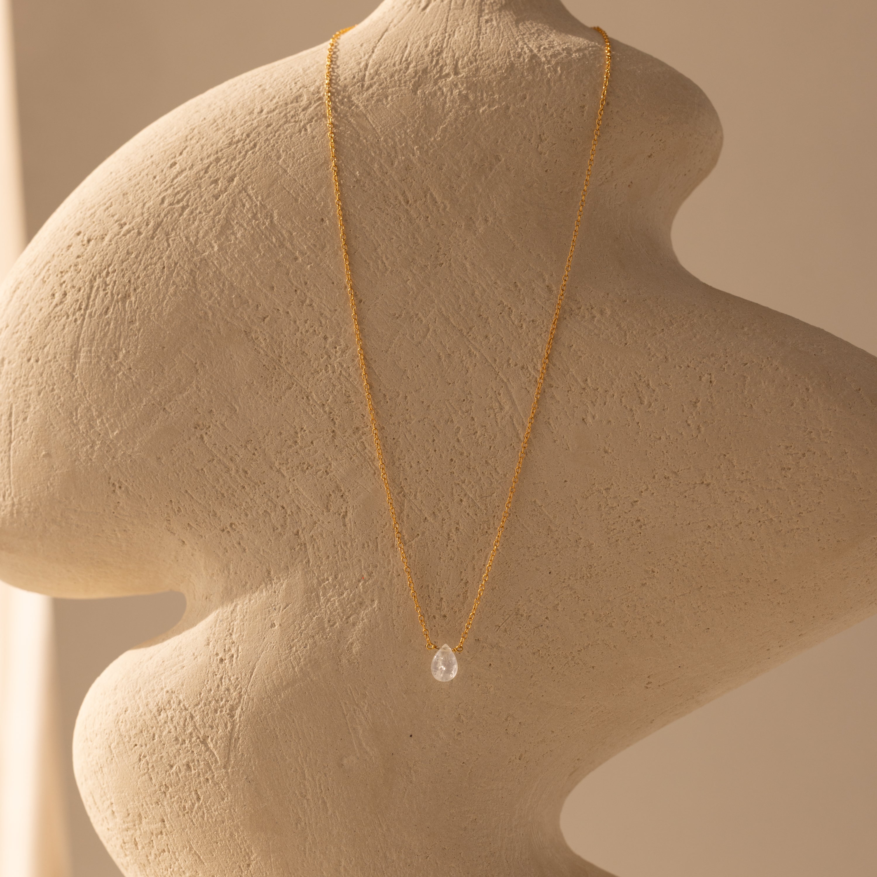 The Teardrop Moonstone Necklace in 18K Gold features a clear moonstone pendant on a gold chain, beautifully displayed on a textured beige abstract sculpture.