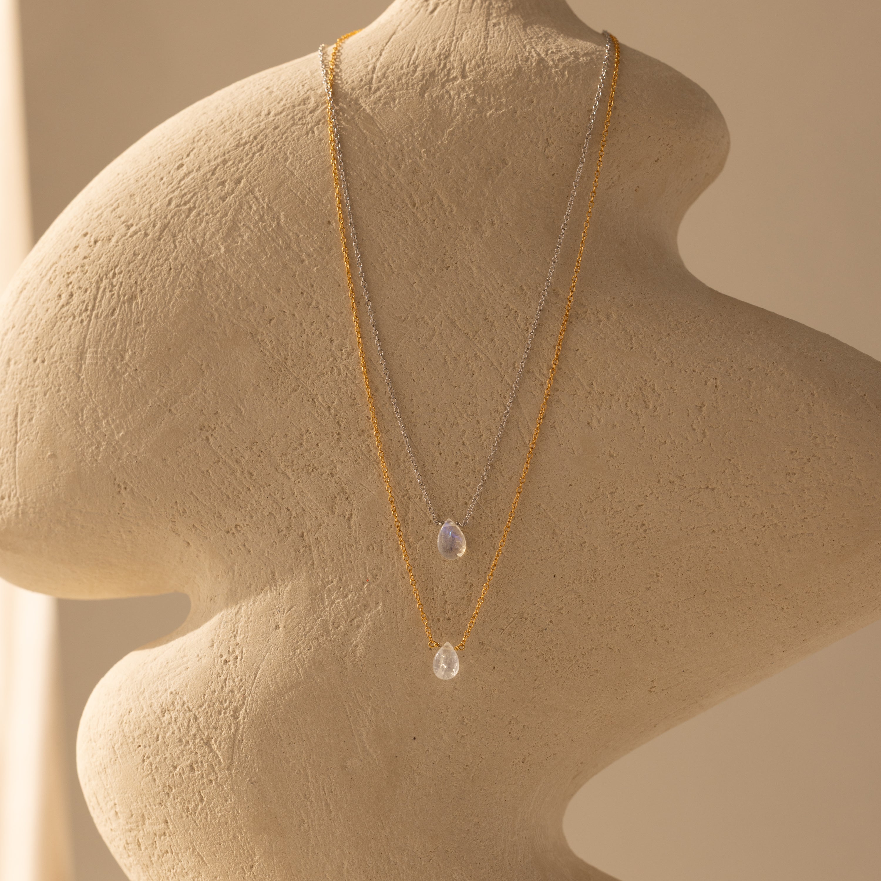 The Teardrop Moonstone Necklace, a delicate minimalist piece with a small pendant, is displayed on a textured, abstract beige mannequin bust.