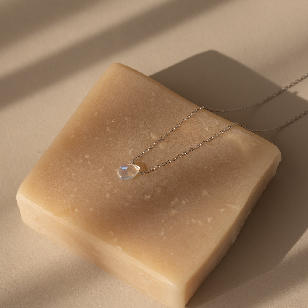 The Teardrop Moonstone Necklace in Sterling Silver is showcased on a beige bar of soap in soft sunlight, embodying the essence of minimalist jewelry.