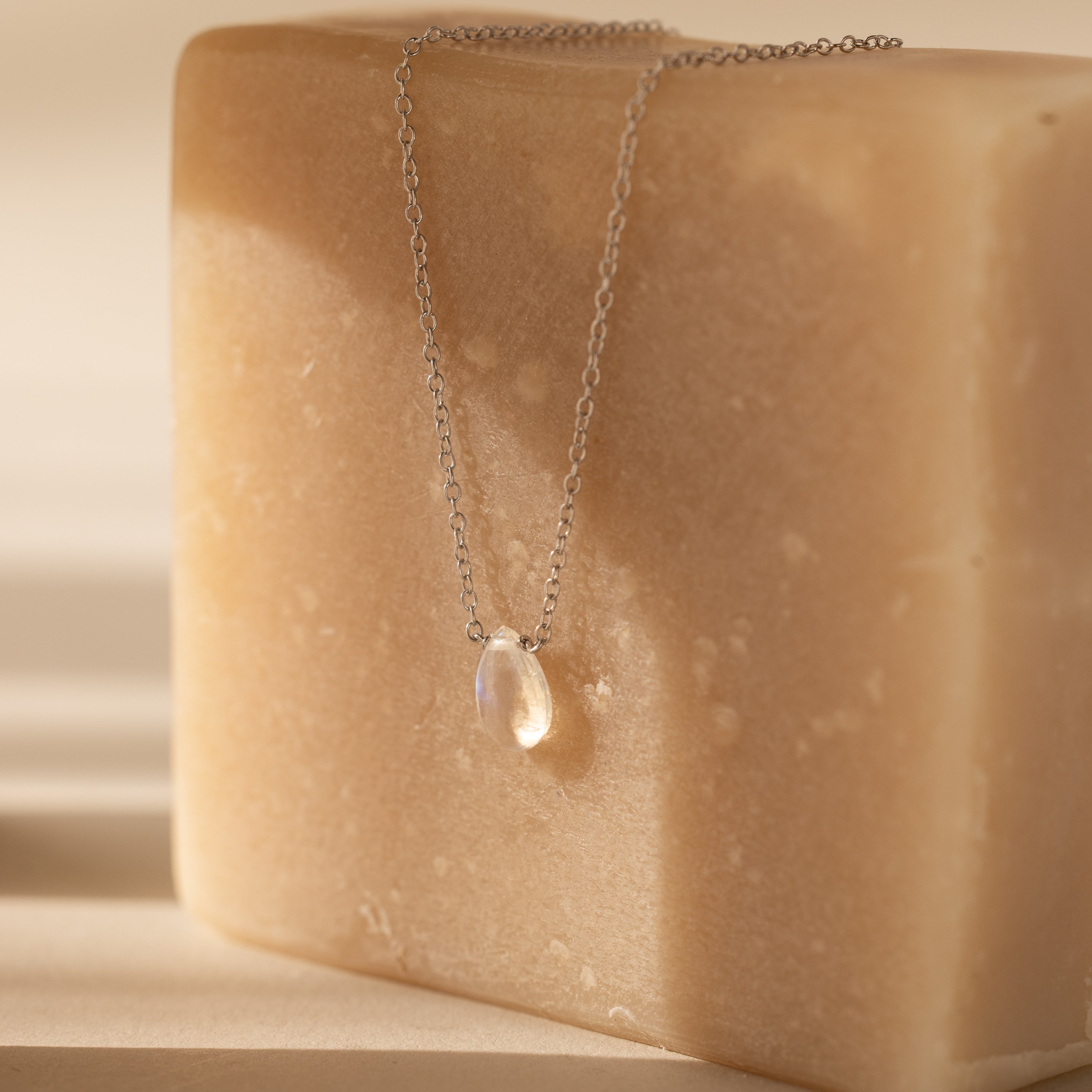The Teardrop Moonstone Necklace in Sterling Silver, featuring a clear moonstone pendant, rests on a large beige soap bar under soft natural light.