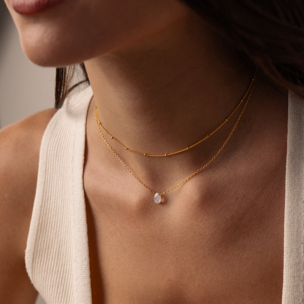 Close-up of a woman wearing the Teardrop Moonstone Necklace in 18K Gold, layered with another gold necklace and paired with a cream-colored sleeveless top.