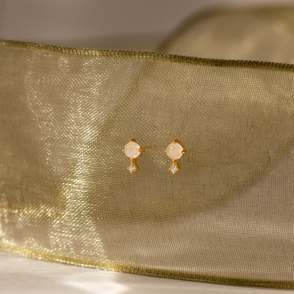 Moonstone Orbit Studs in 18K Gold feature round moonstone studs and delicate dangling crystals, showcased against sheer gold fabric for an elegant touch.