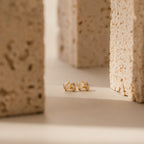 Moonstone Orbit Studs in 18K Gold are displayed on a beige surface between textured stone blocks, emphasizing the timeless elegance of gold and the delicate beauty of moonstone.