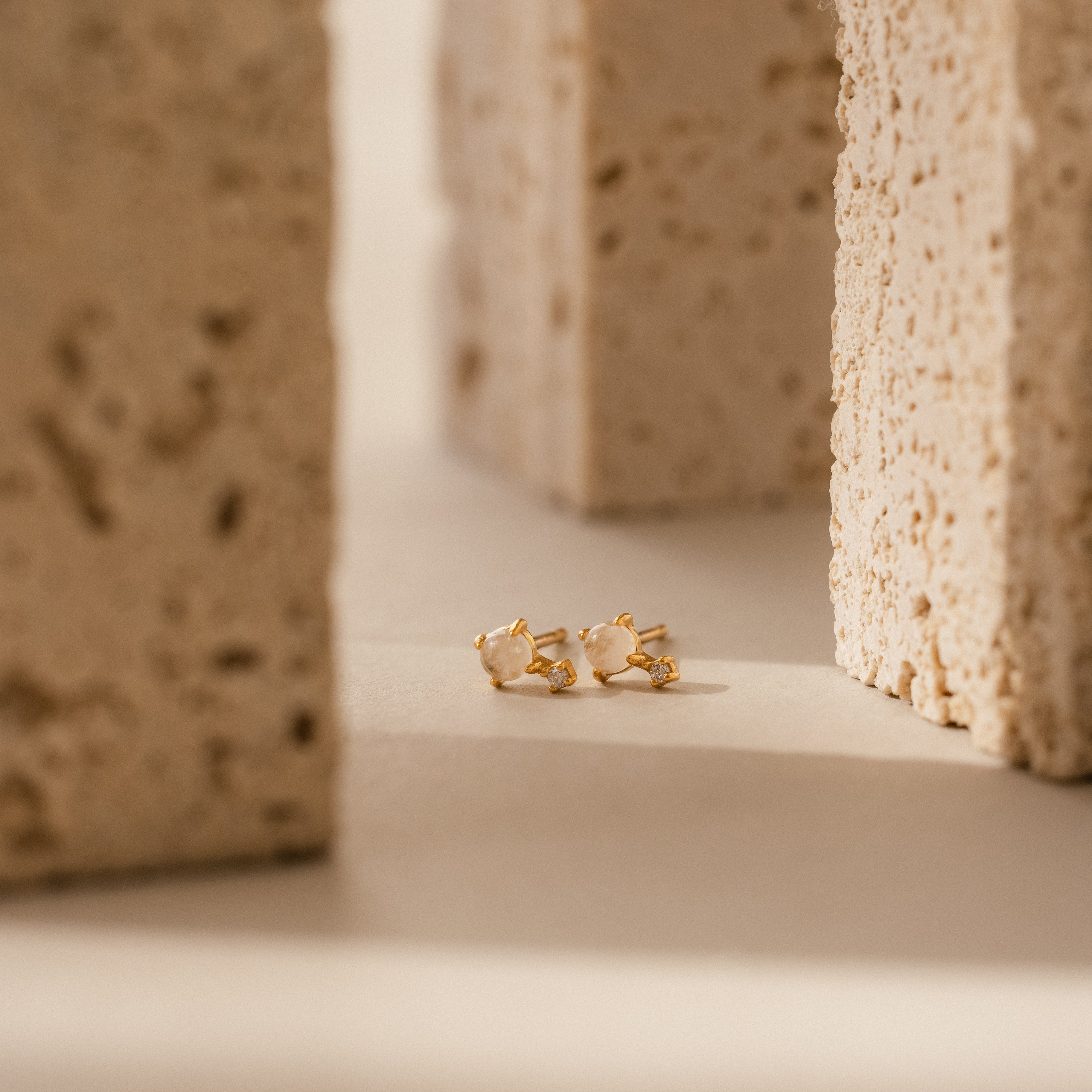 Moonstone Orbit Studs rest on a beige surface between rough stone blocks, showcasing timeless elegance.