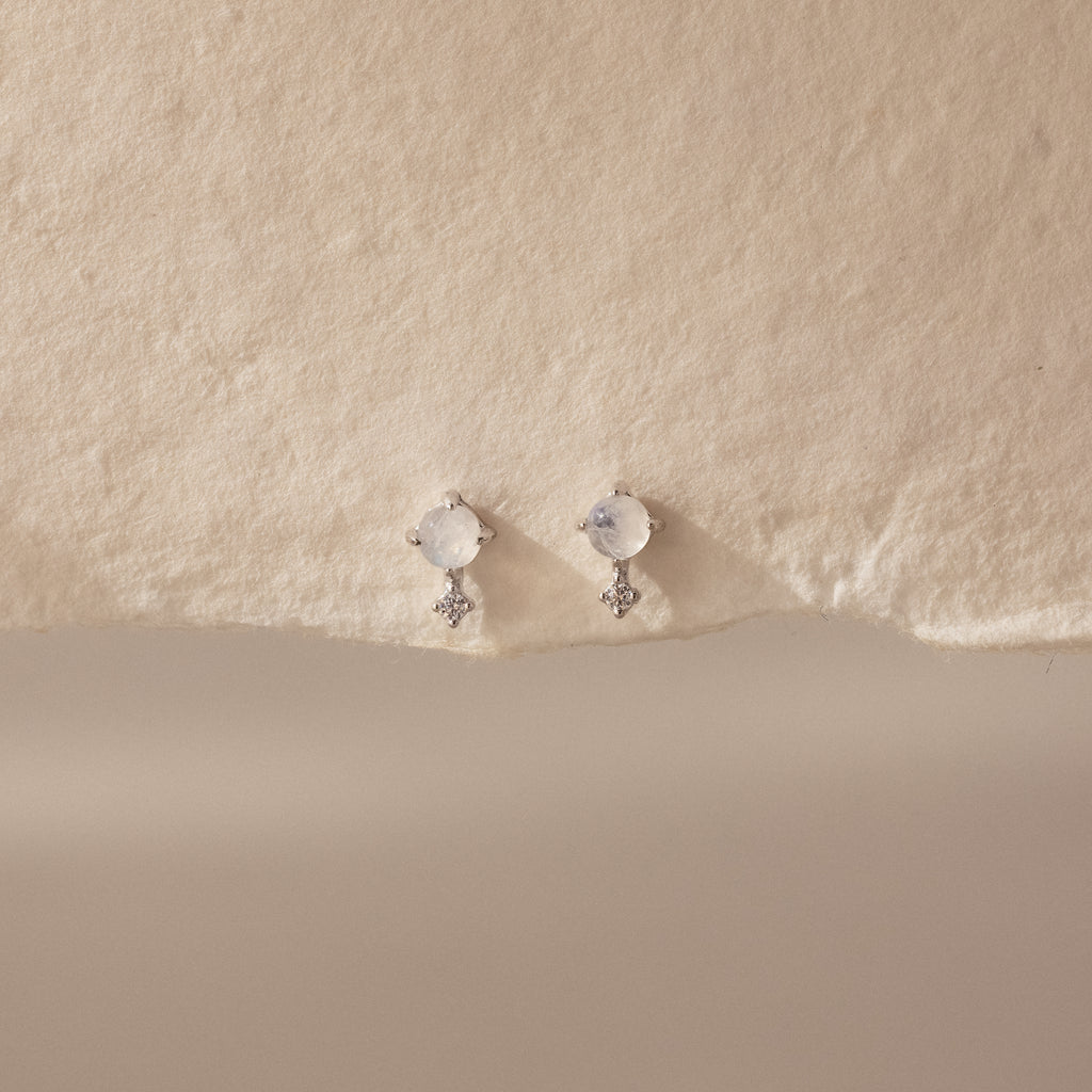 The Moonstone Orbit Studs feature delicate gold with round moonstones and small dangling crystals, creating a subtle sparkle on a textured cream background.