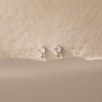 The Moonstone Orbit Studs feature delicate gold with round moonstones and small dangling crystals, creating a subtle sparkle on a textured cream background.