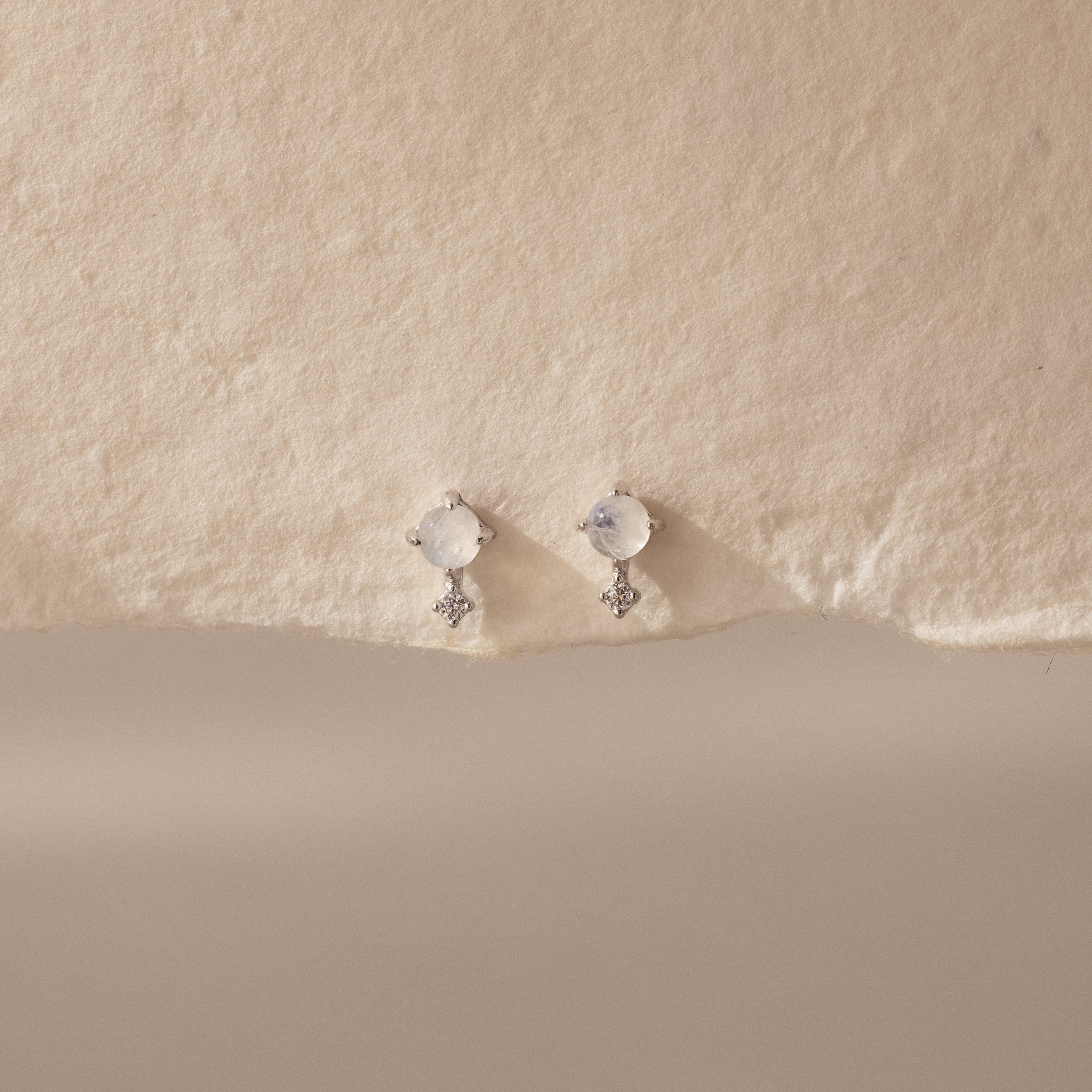 The Moonstone Orbit Studs feature delicate gold with round moonstones and small dangling crystals, creating a subtle sparkle on a textured cream background.