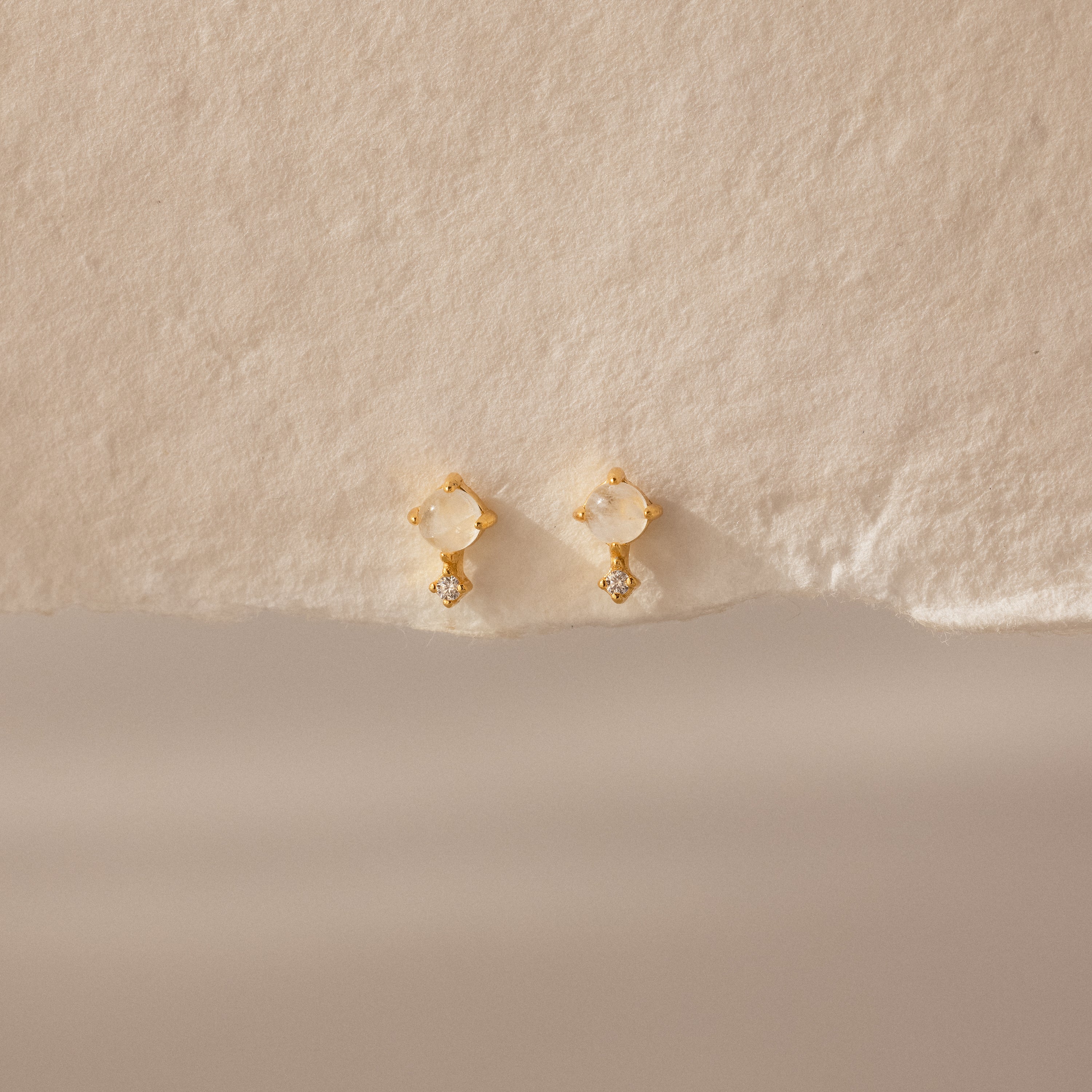 Moonstone Orbit Studs featuring gold, pearl studs, and small diamonds are displayed on textured beige paper. These diamond accent earrings add a touch of elegance to any look.