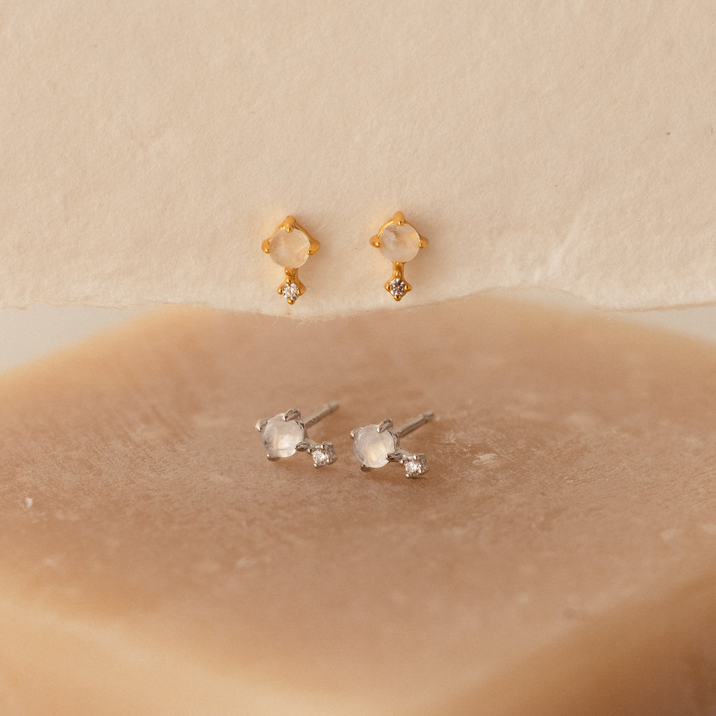 A pair of Moonstone Orbit Studs is displayed on a beige surface alongside classic silver diamond accent earrings, highlighting the elegant design of gold moonstone studs and timeless silver accents.