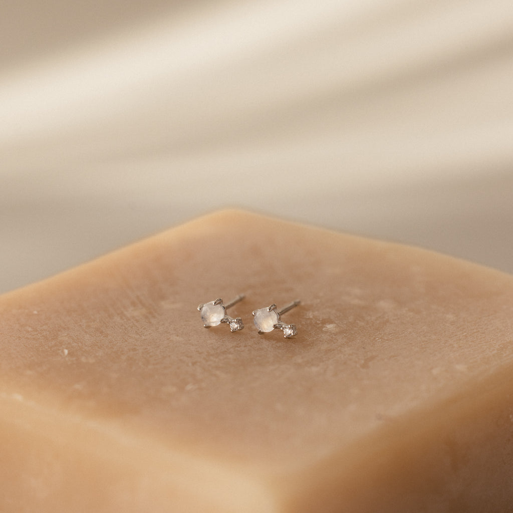 A pair of Moonstone Orbit Studs in sterling silver with small white stones displayed on a beige textured block.