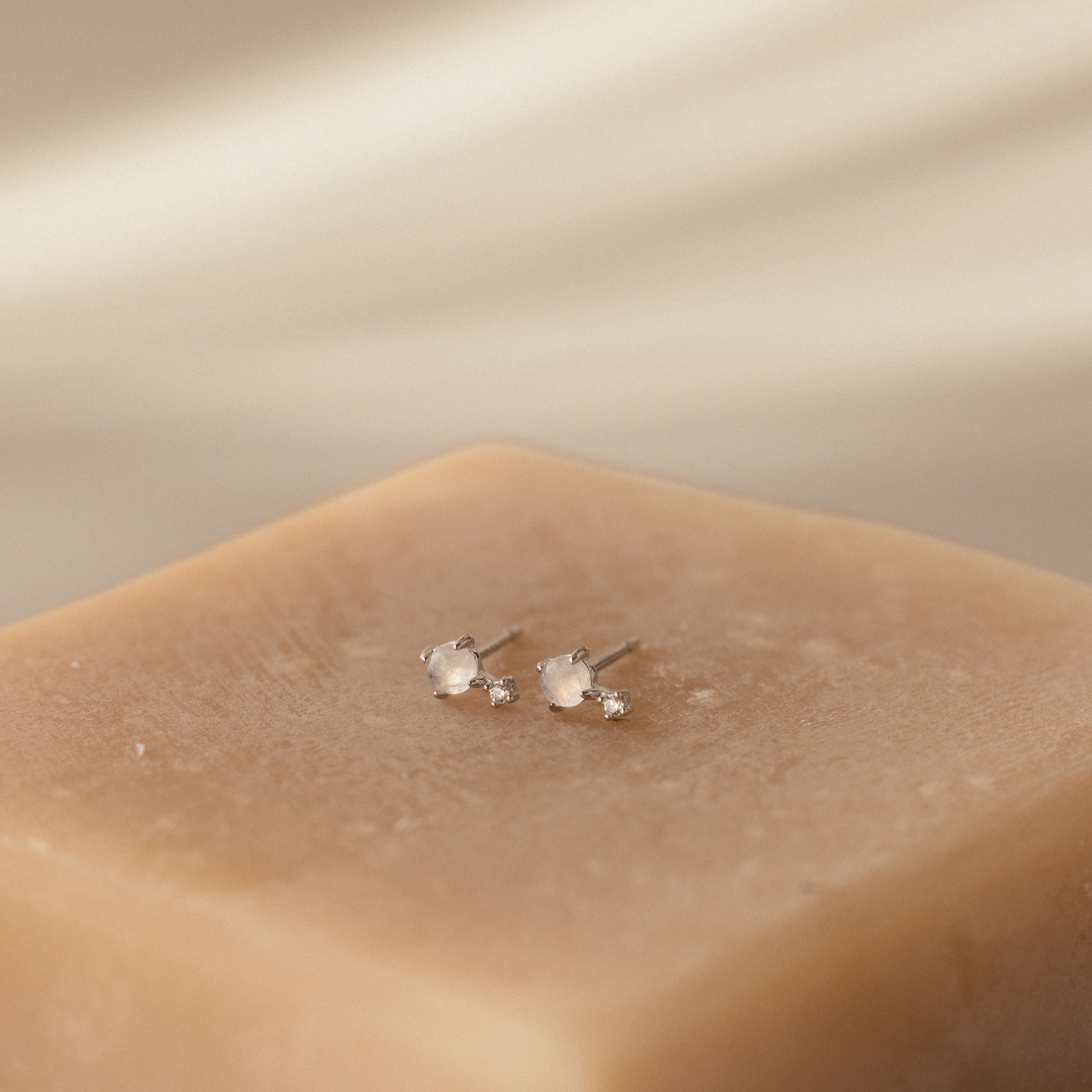 A pair of Moonstone Orbit Studs in sterling silver with small white stones displayed on a beige textured block.