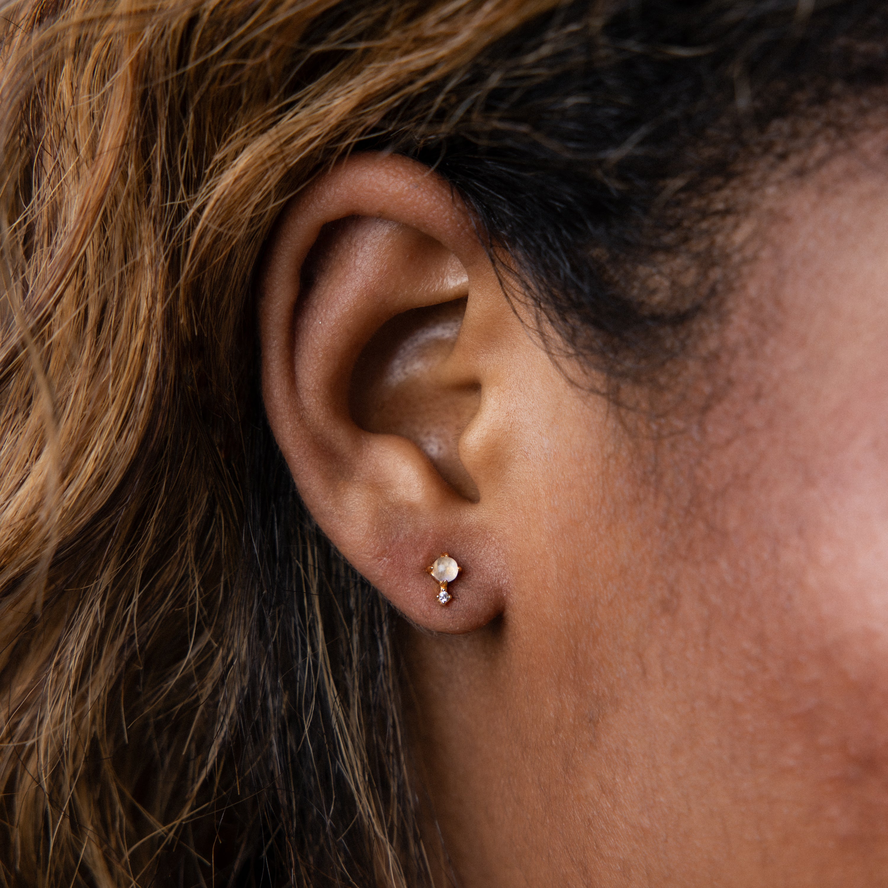 Close-up of an ear wearing Moonstone Orbit Studs—small gold earrings with moonstone accents—brown hair partially visible. A timeless, elegant choice for those who appreciate classic gold jewelry with a unique twist.