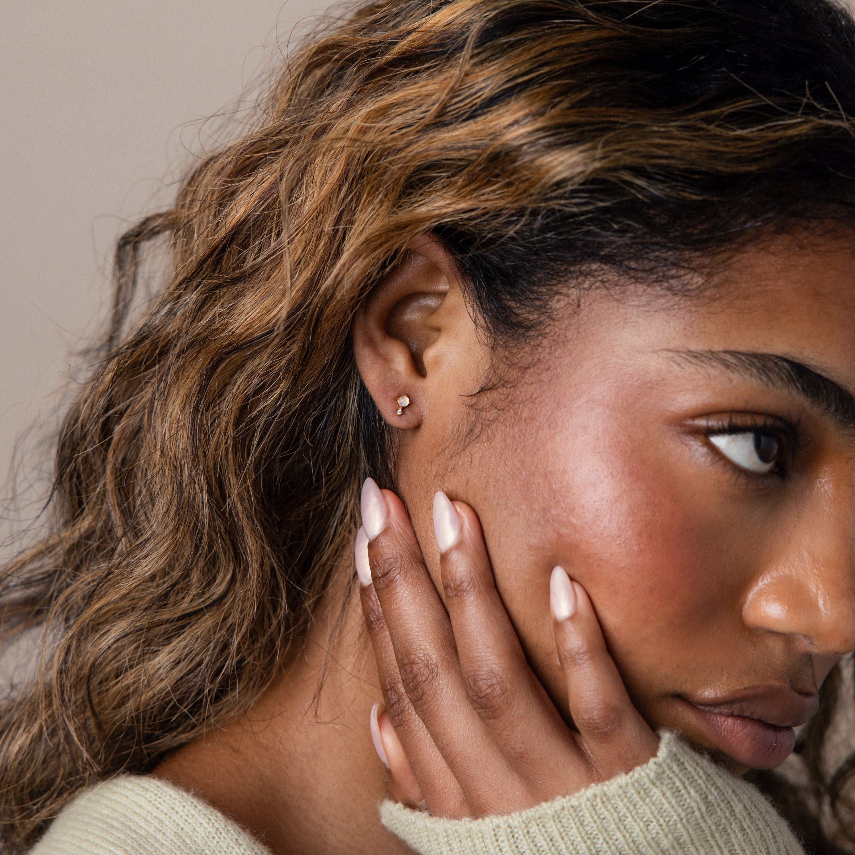 Woman with wavy hair and manicured nails touches her face, wearing elegant Moonstone Orbit Studs in 18K gold.