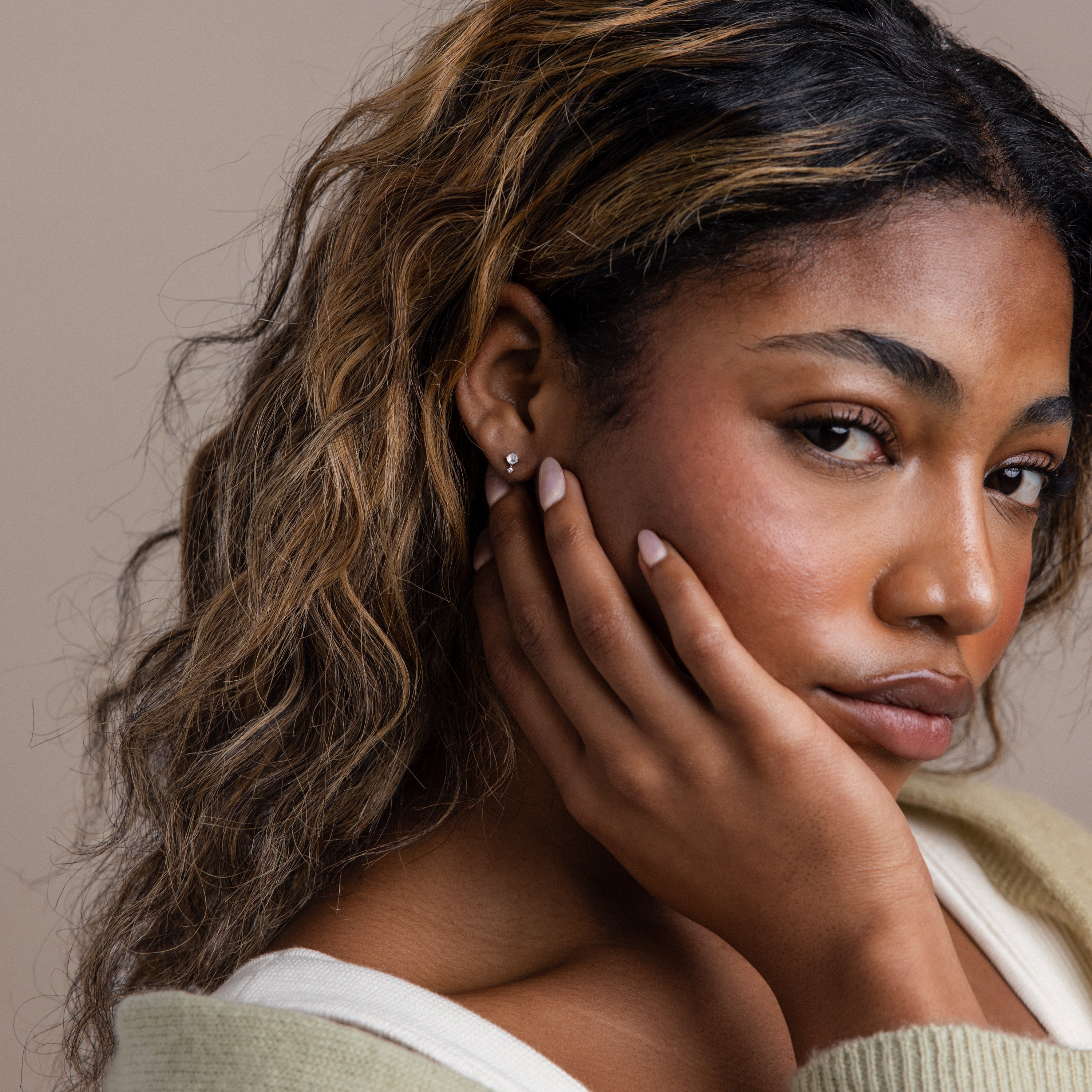 A woman with wavy brown hair rests her face on her hand, gazing thoughtfully at the camera while wearing elegant Moonstone Orbit Studs in Sterling Silver that shimmer in the light.