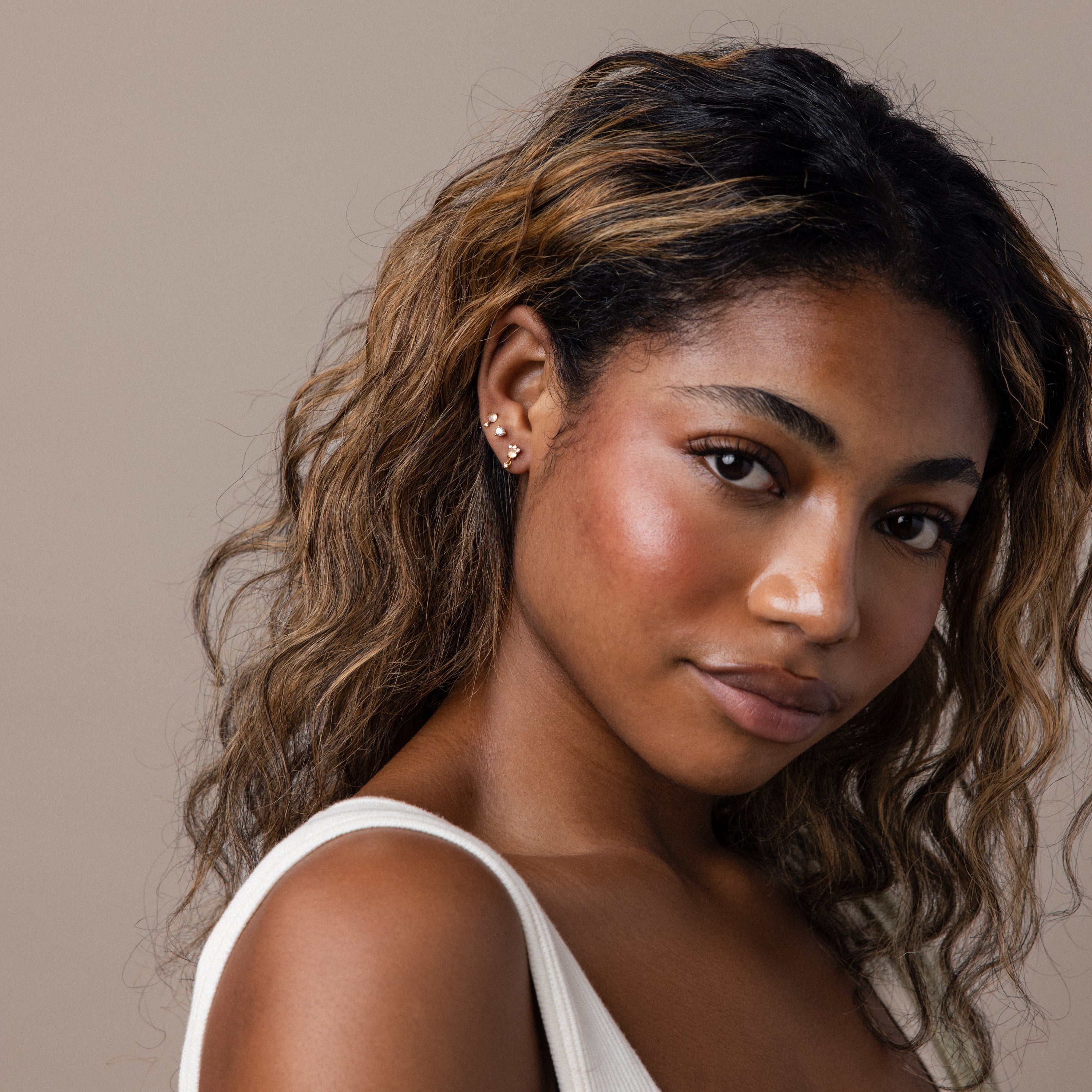Woman with wavy brown hair, wearing a white tank top and the Moonstone Orbit Studs in 18K Gold, looking at the camera.