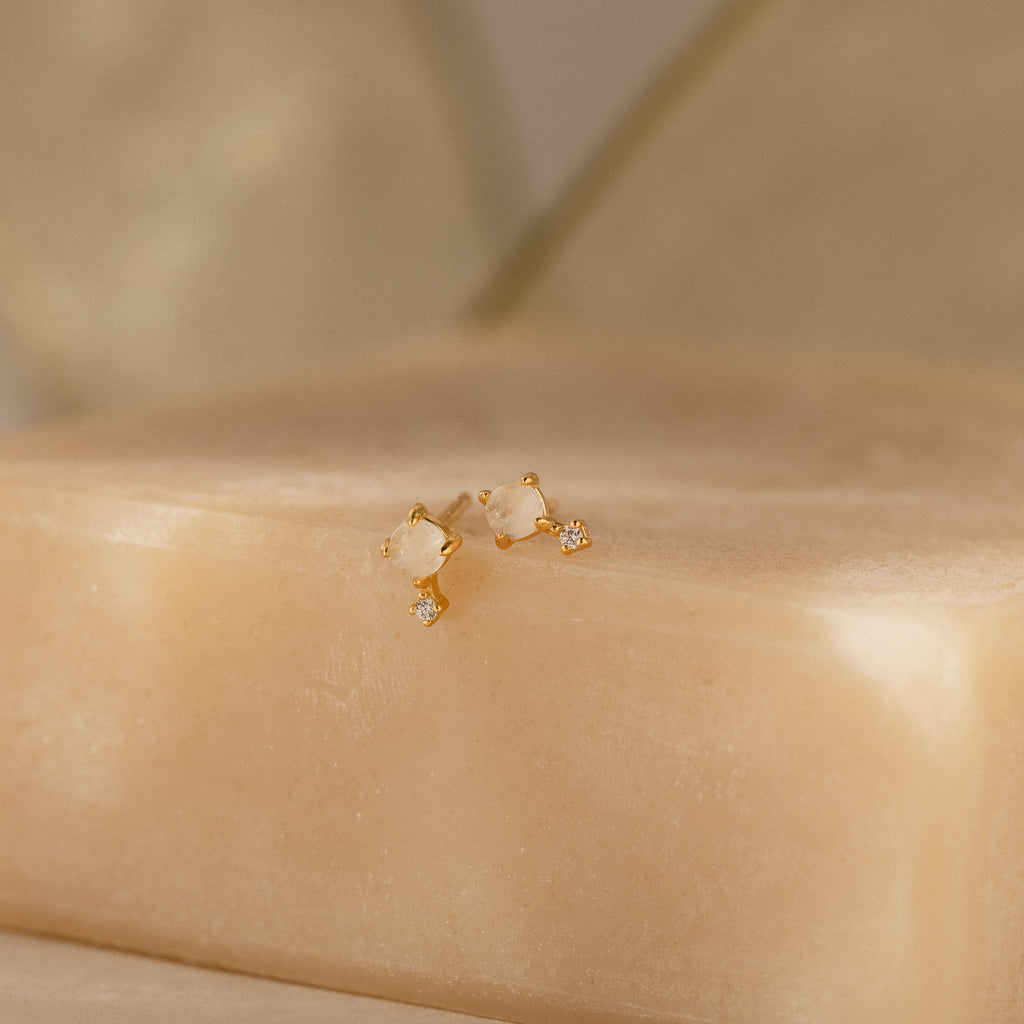 The Moonstone Orbit Studs in 18K Gold are displayed on a beige soap bar with a soft, neutral background.