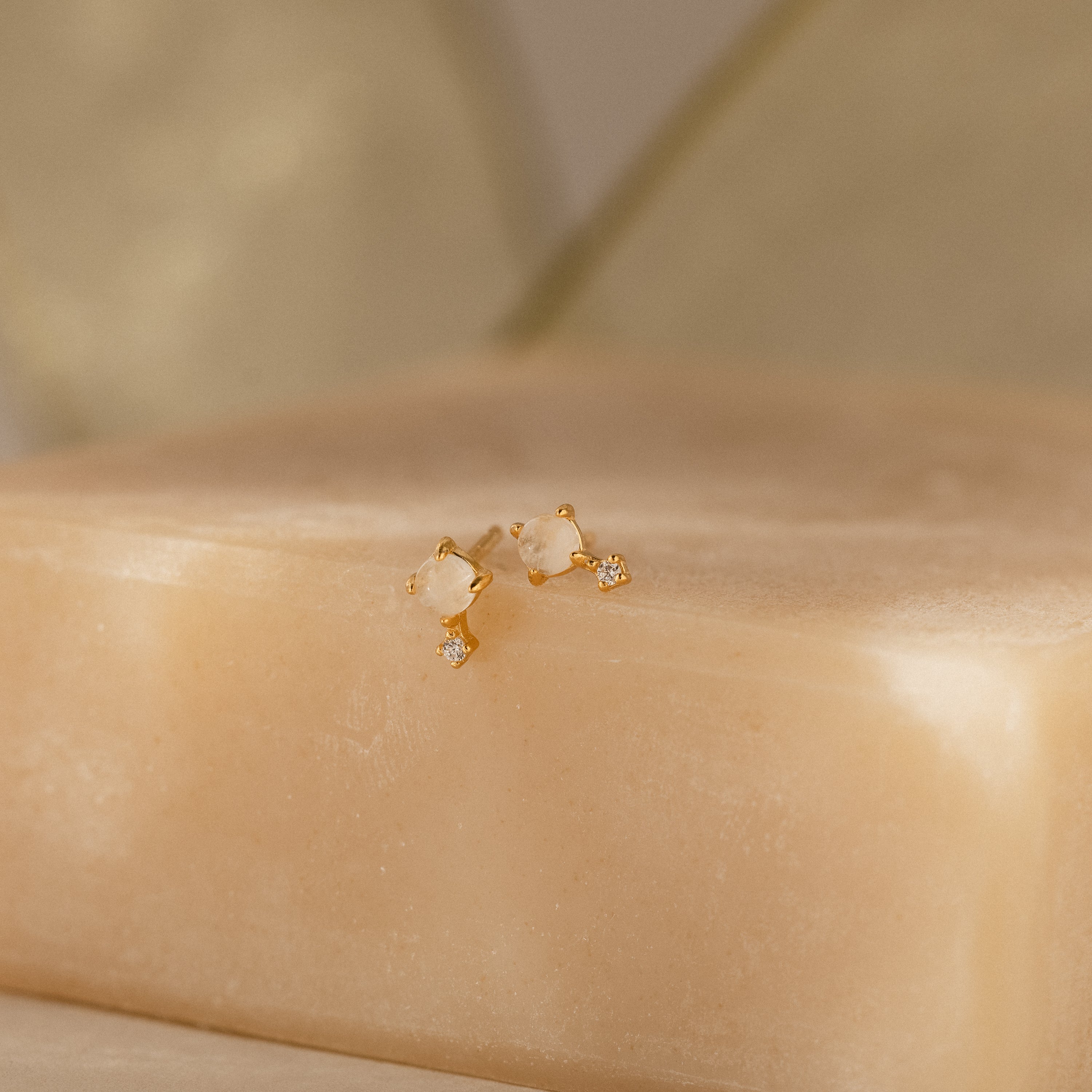 The Moonstone Orbit Studs in 18K Gold are displayed on a beige soap bar with a soft, neutral background.