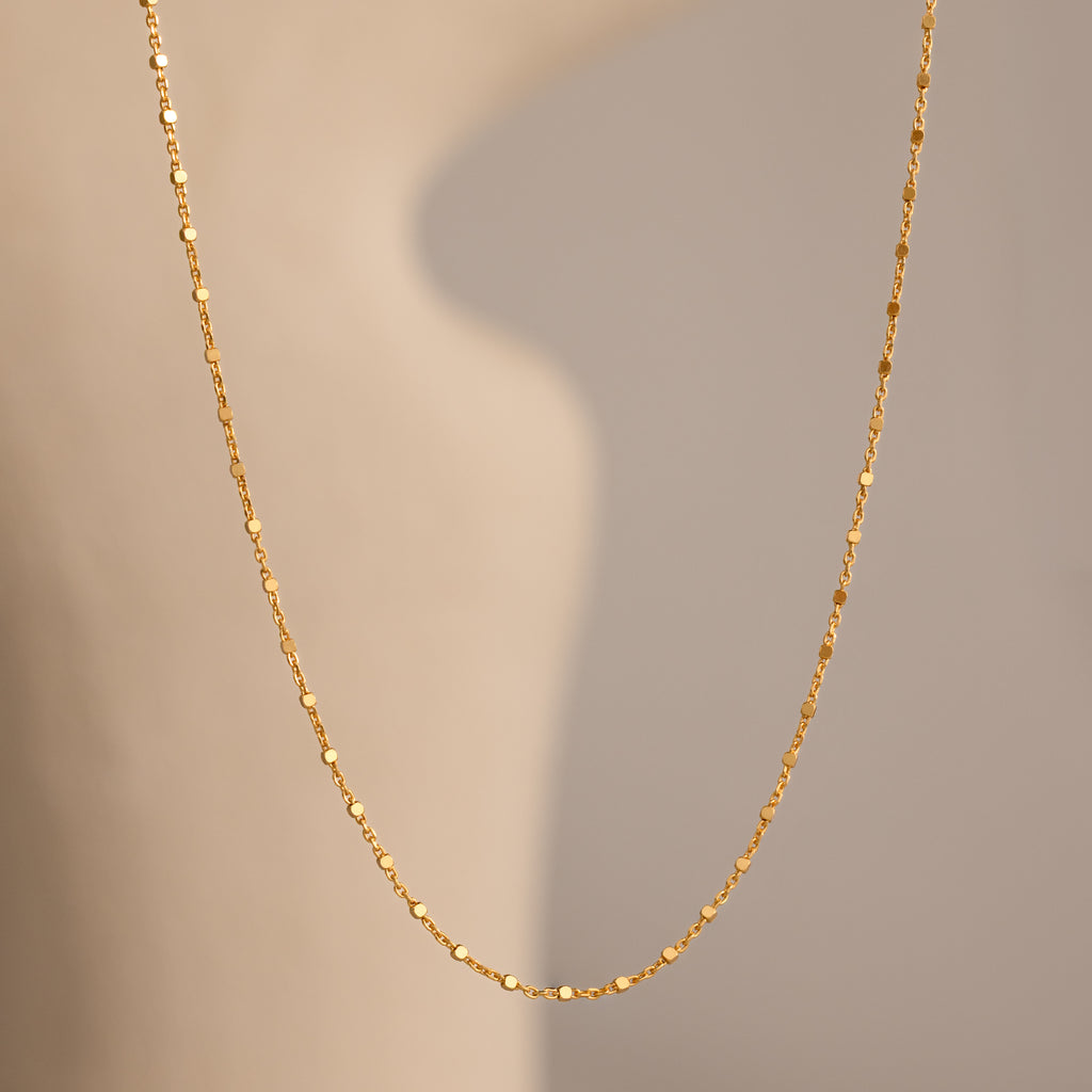 The Dot Station Necklace in 18K Gold hangs gracefully against a soft, beige background—an elegant choice for any minimalist jewelry lover.