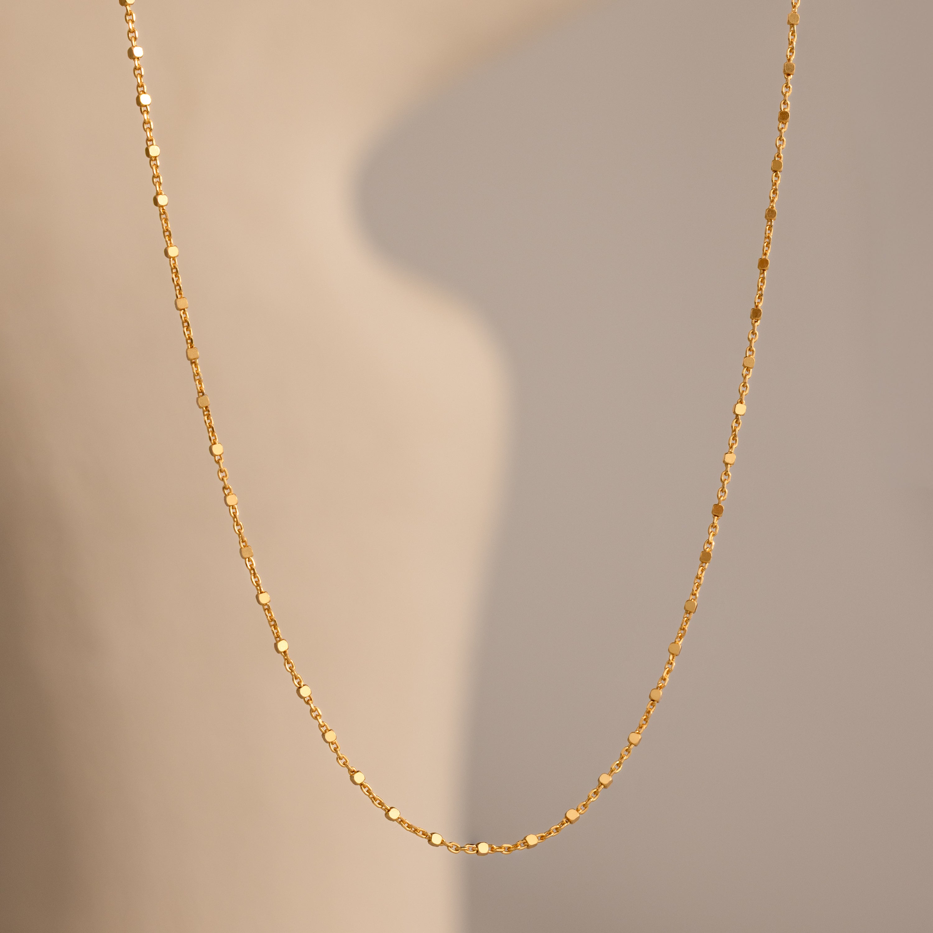 The Dot Station Necklace in 18K Gold hangs gracefully against a soft, beige background—an elegant choice for any minimalist jewelry lover.