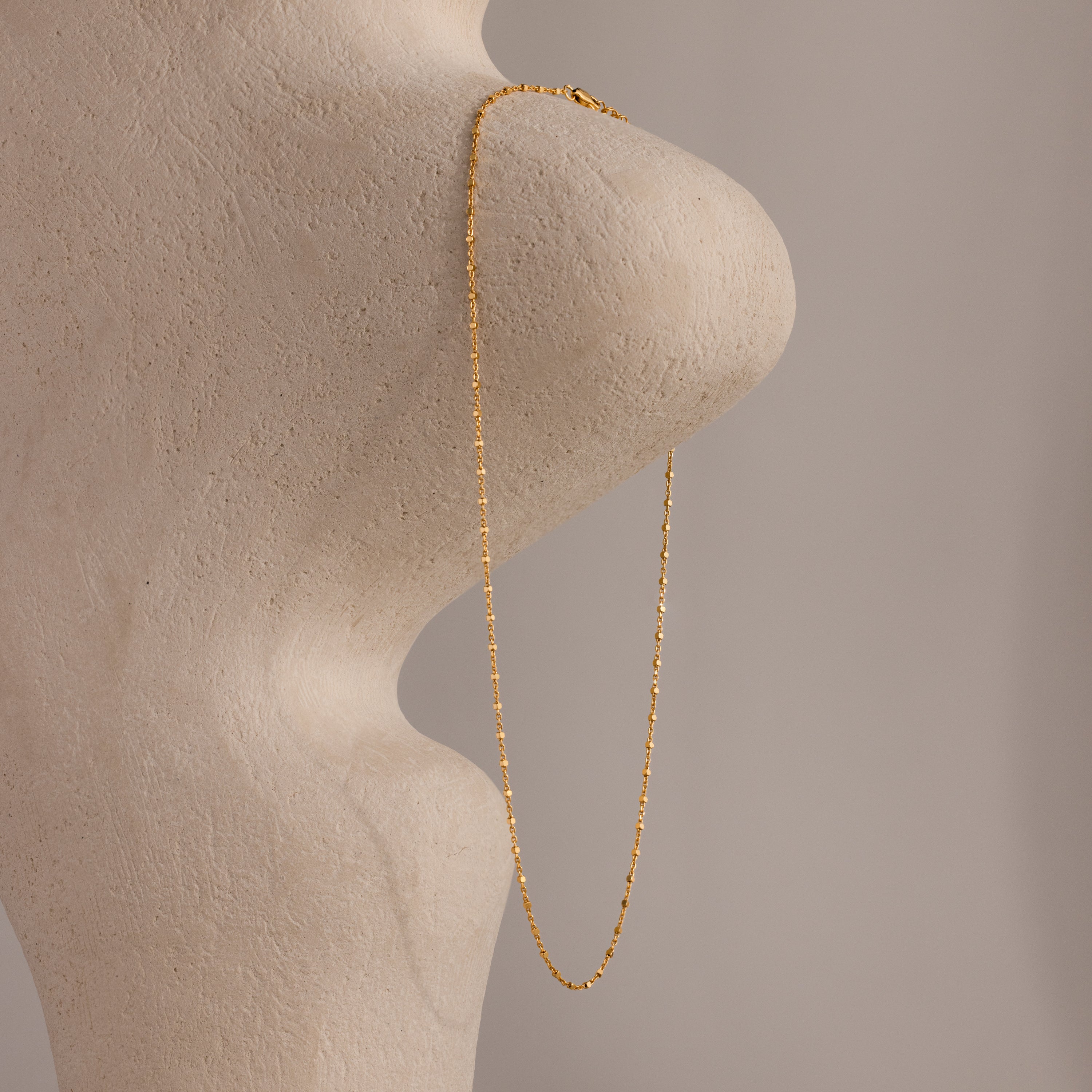 The Dot Station Necklace in 18K Gold is draped over a textured, abstract beige sculpture set against a neutral background.