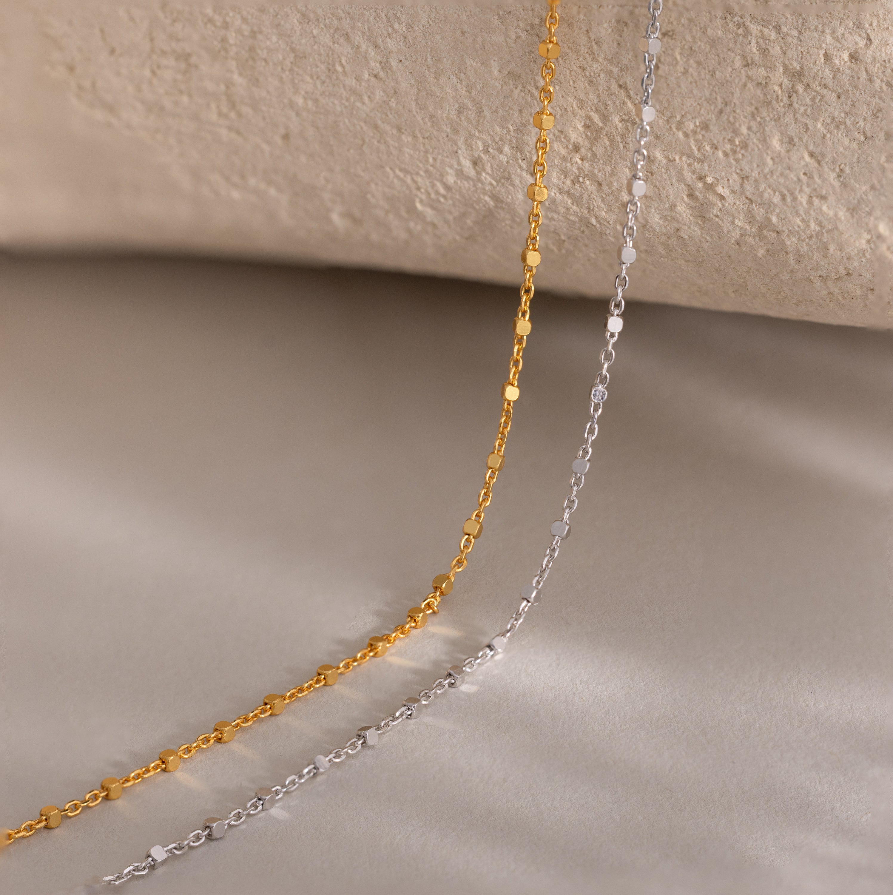 Two delicate Dot Station Necklaces, one gold and one silver, rest on a soft beige surface with a textured background—ideal for those who love minimalist necklace styles.