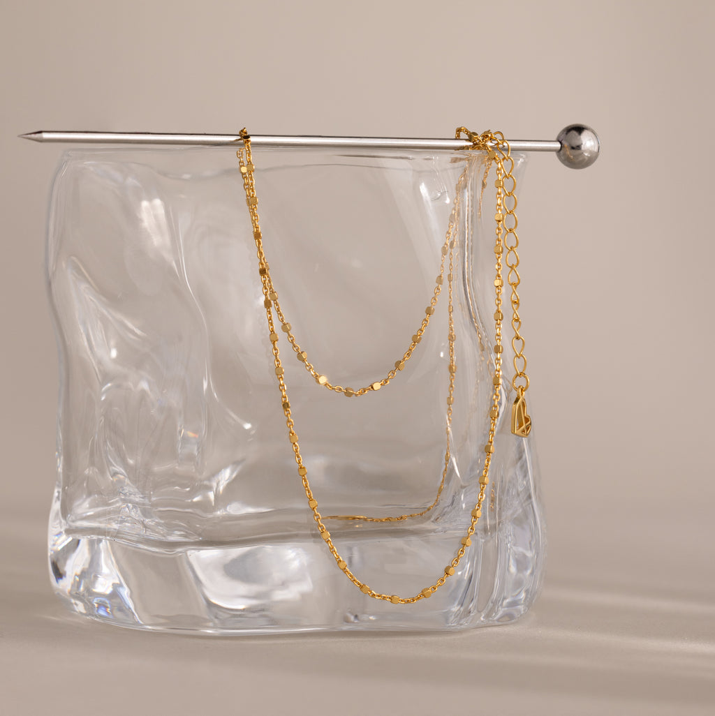 A delicate chain from the Dot Station Necklace in 18K Gold is draped over a glass block, with a silver pin resting on top.