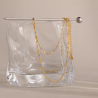 A delicate chain from the Dot Station Necklace in 18K Gold is draped over a glass block, with a silver pin resting on top.