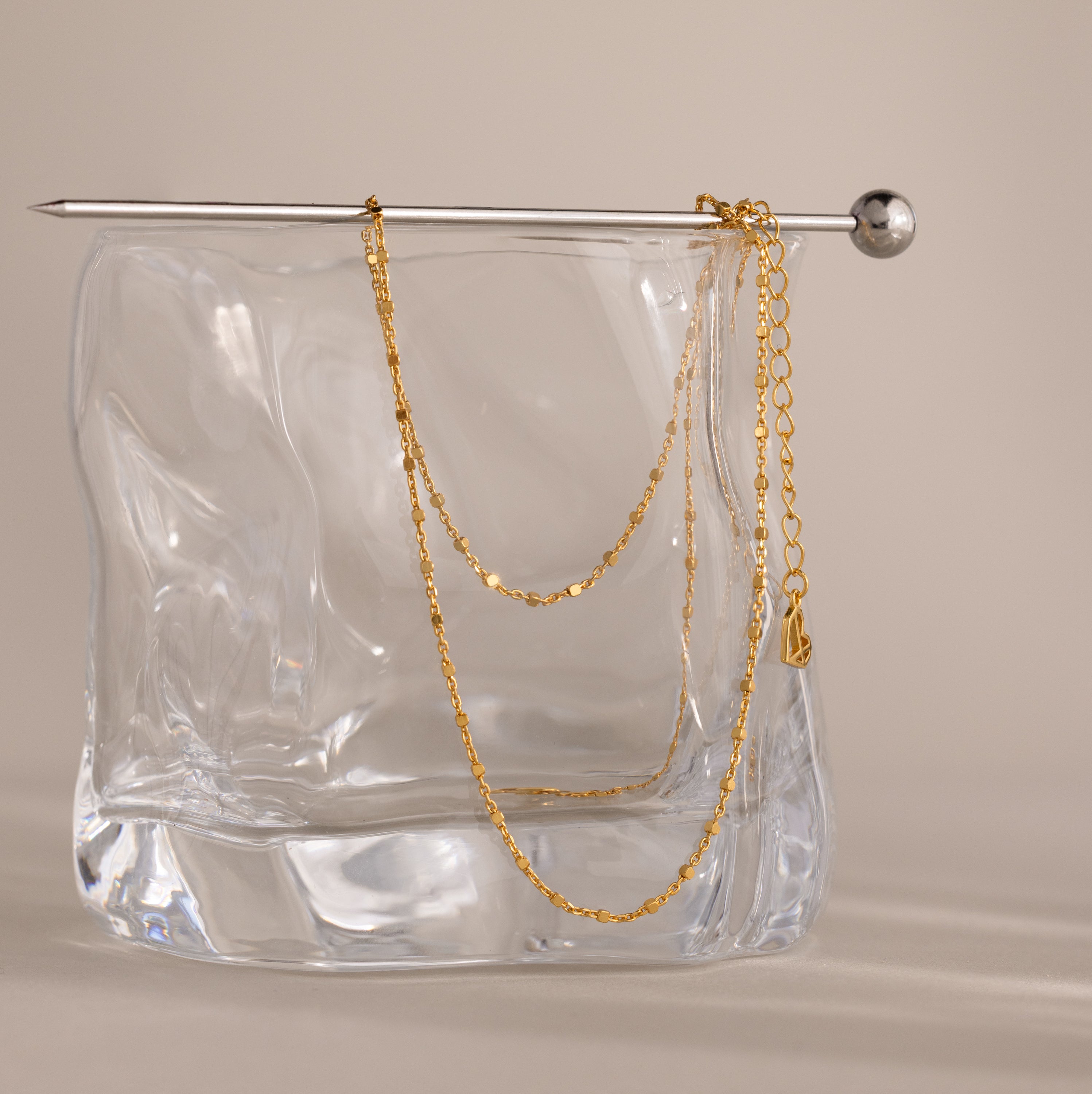 A delicate chain from the Dot Station Necklace in 18K Gold is draped over a glass block, with a silver pin resting on top.