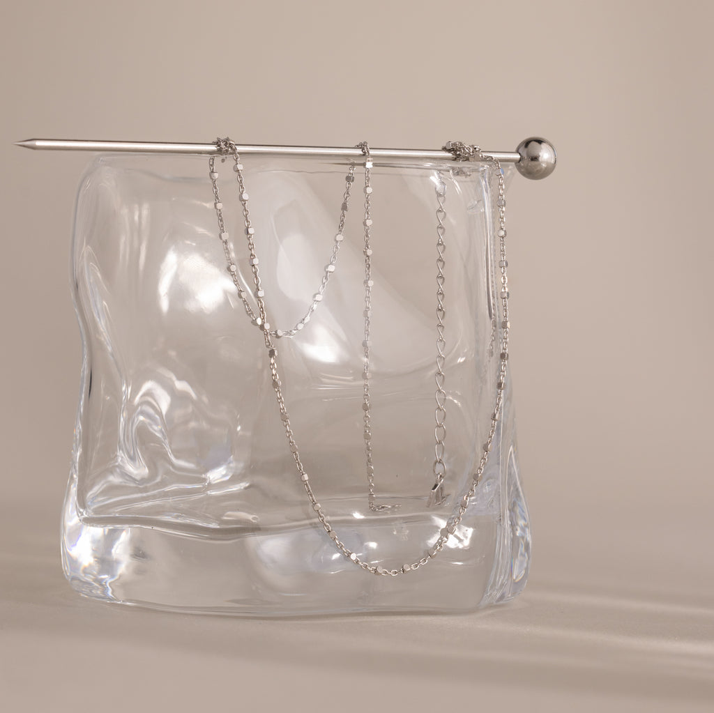 A delicate Dot Station Necklace in Sterling Silver is draped over a clear, wavy glass block with a silver rod on top, highlighting the minimalist piece’s refined elegance.