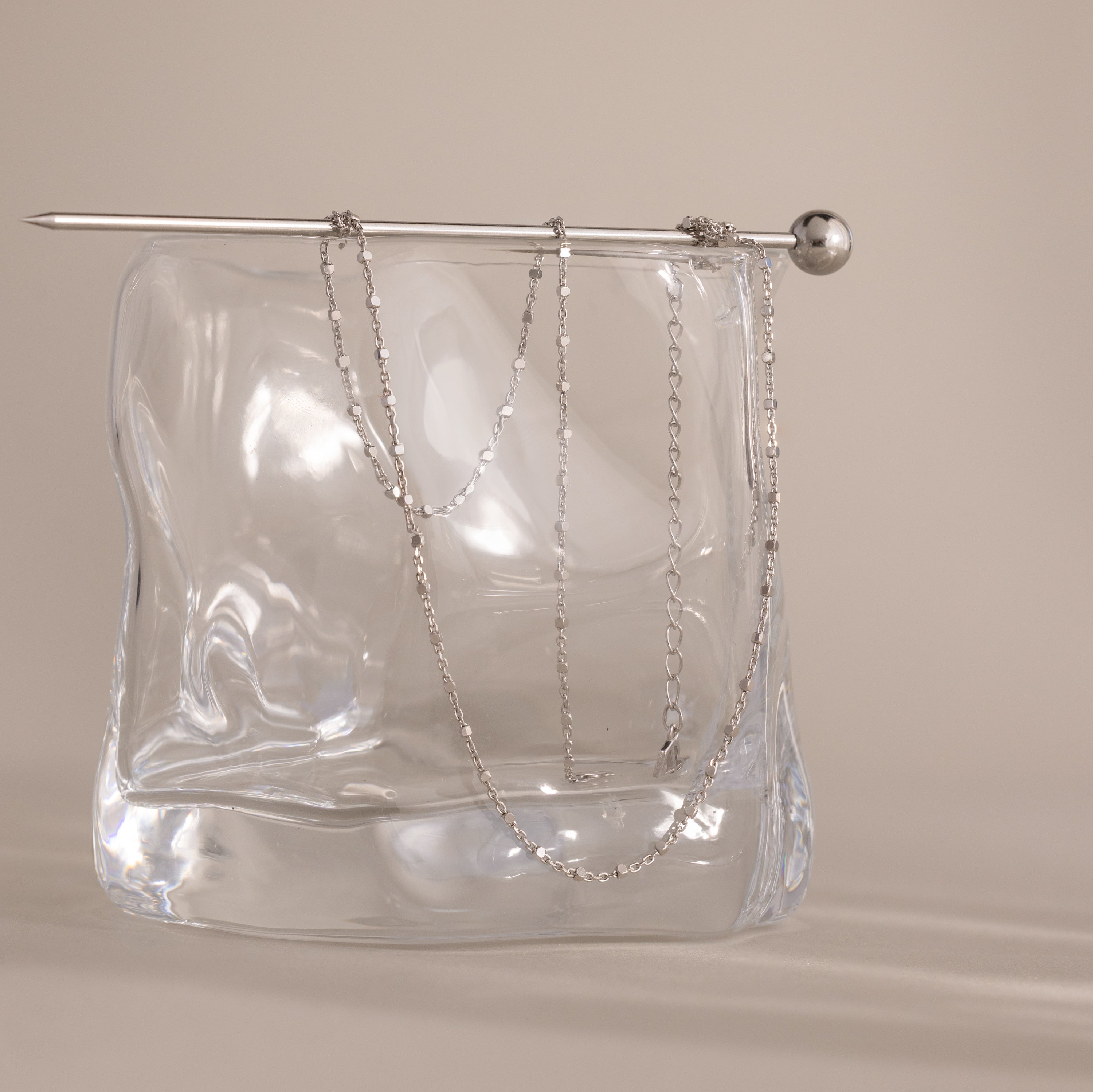 A delicate Dot Station Necklace in Sterling Silver is draped over a clear, wavy glass block with a silver rod on top, highlighting the minimalist piece’s refined elegance.