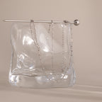 The Dot Station Necklace, a delicate minimalist chain, is draped over a clear wavy glass block with a silver rod resting on top.