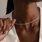 A person wearing the Dot Station Necklace in 18K Gold with a delicate chain and a cream-colored top, gently holding the necklace.