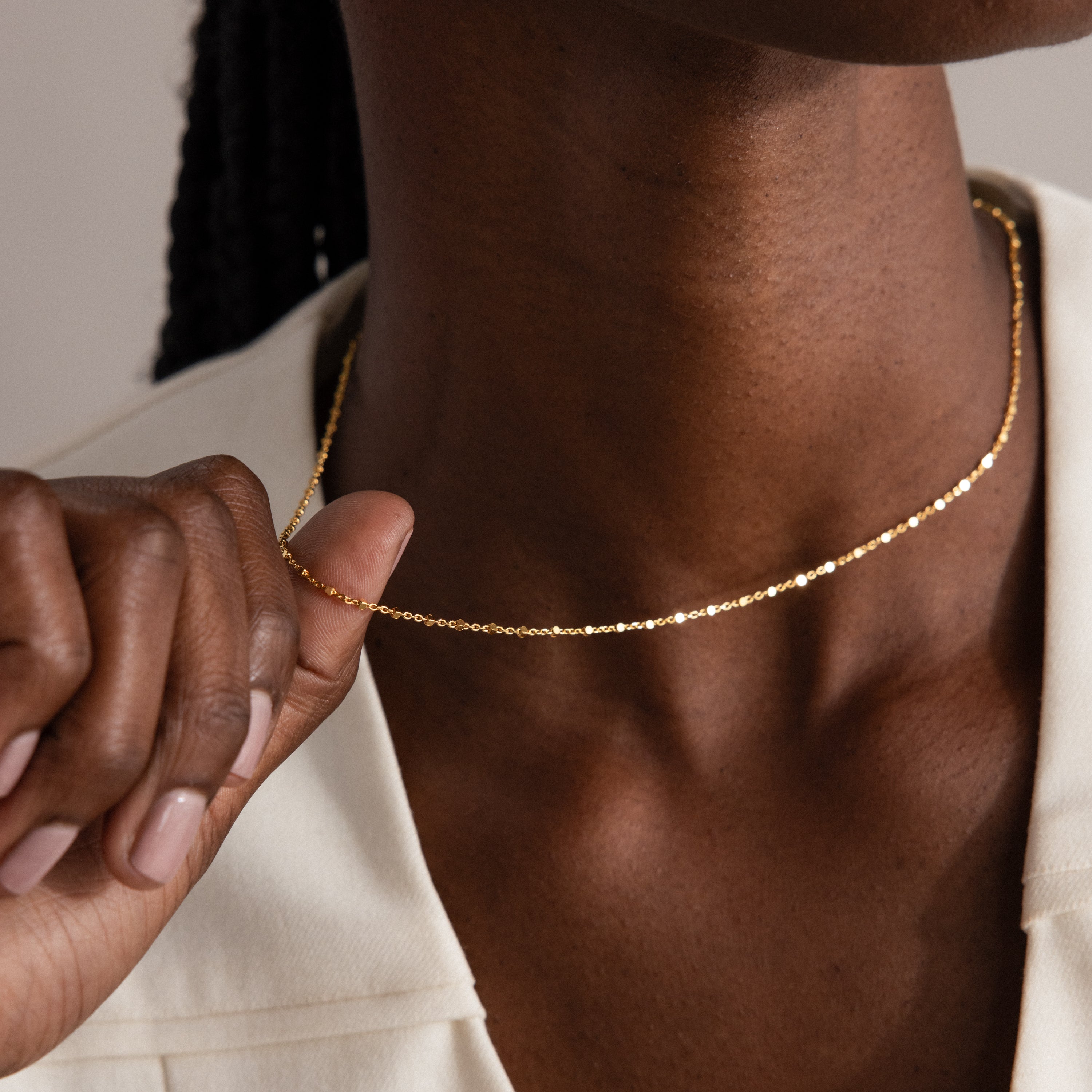 Dressed in cream, a person holds the Dot Station Necklace—a delicate, minimalist chain—near their neck.