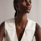 A woman wearing a cream sleeveless top and layered gold necklaces, including a delicate chain and Dot Station Necklace in 18K Gold, gazes upwards.